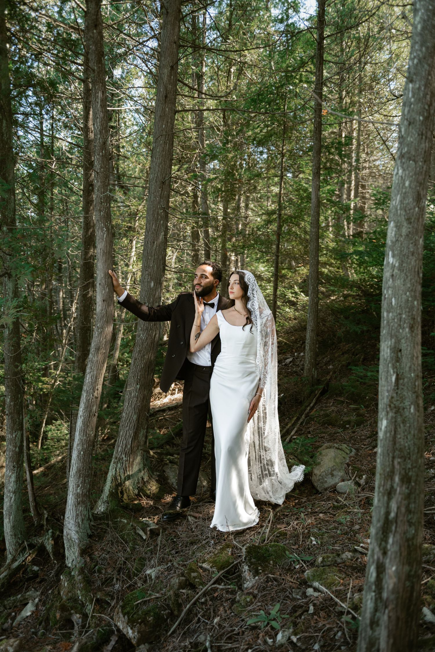 Bride in white gown with lace veil and groom in tuxedo standing among tall trees in a sunlit forest.