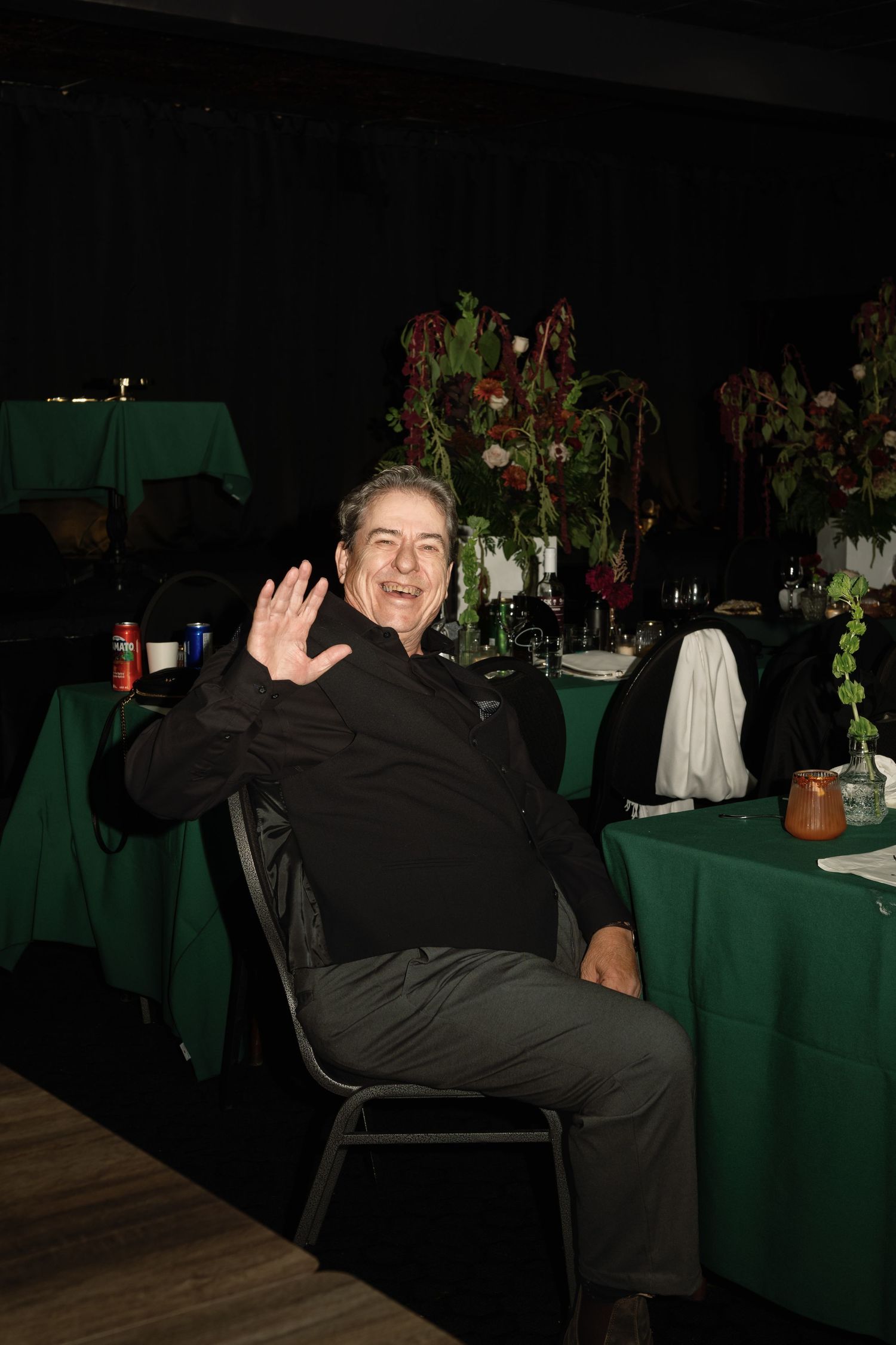 A smiling man waving at a formal event, seated at a green-draped table with floral centerpieces in a dark venue.
