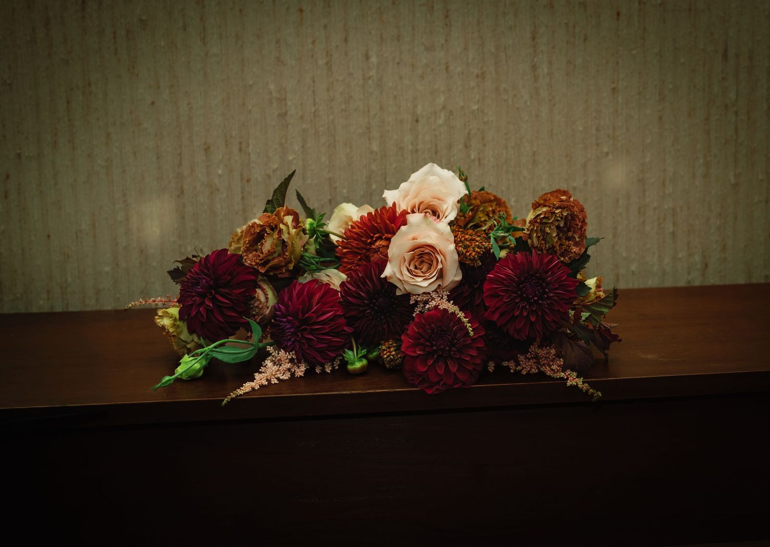 Elegant floral arrangement with deep red dahlias, blush roses, and burgundy blooms displayed on a wooden surface.
