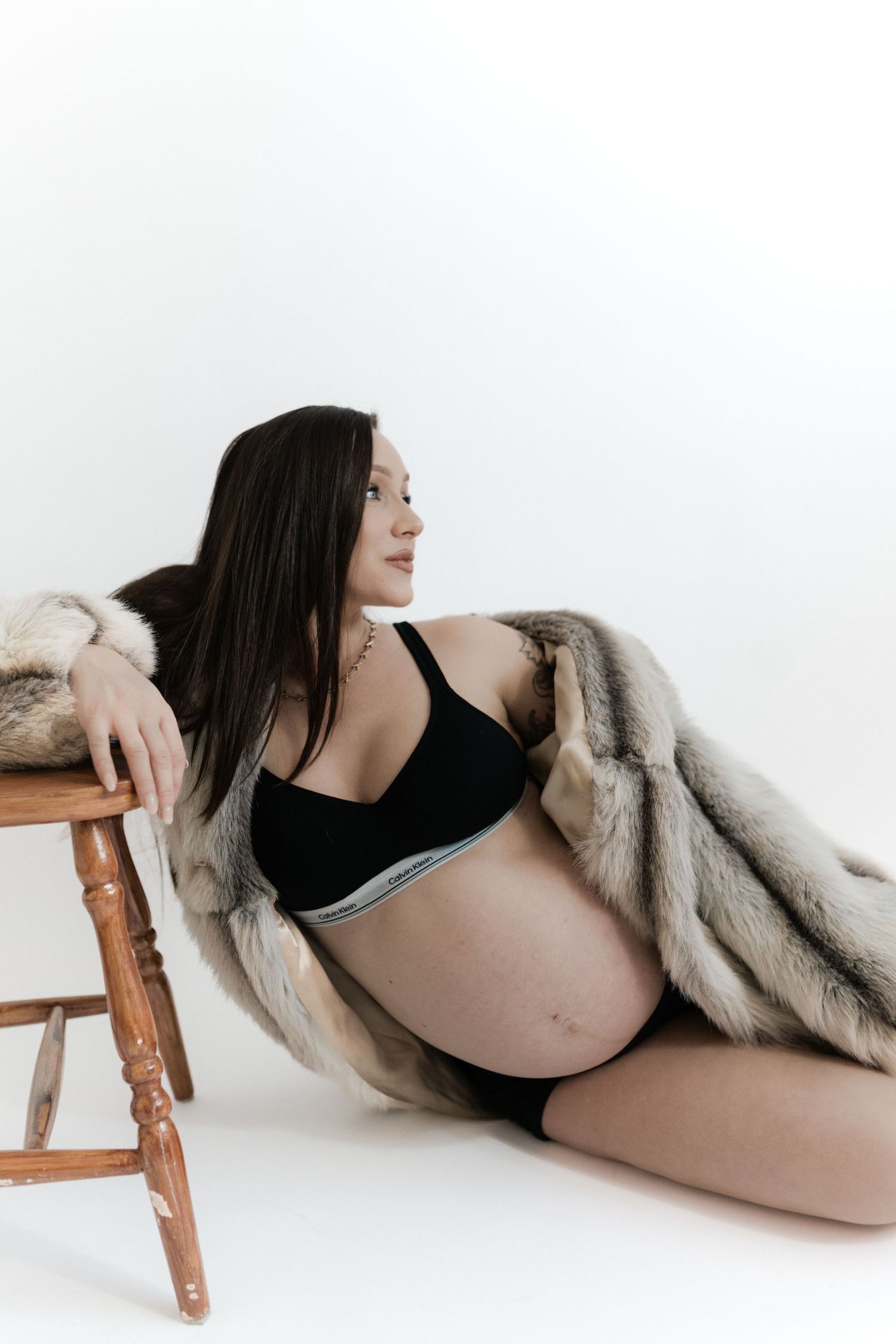 Pregnant woman in black lingerie and fur coat sitting on floor leaning on wooden stool in white studio.