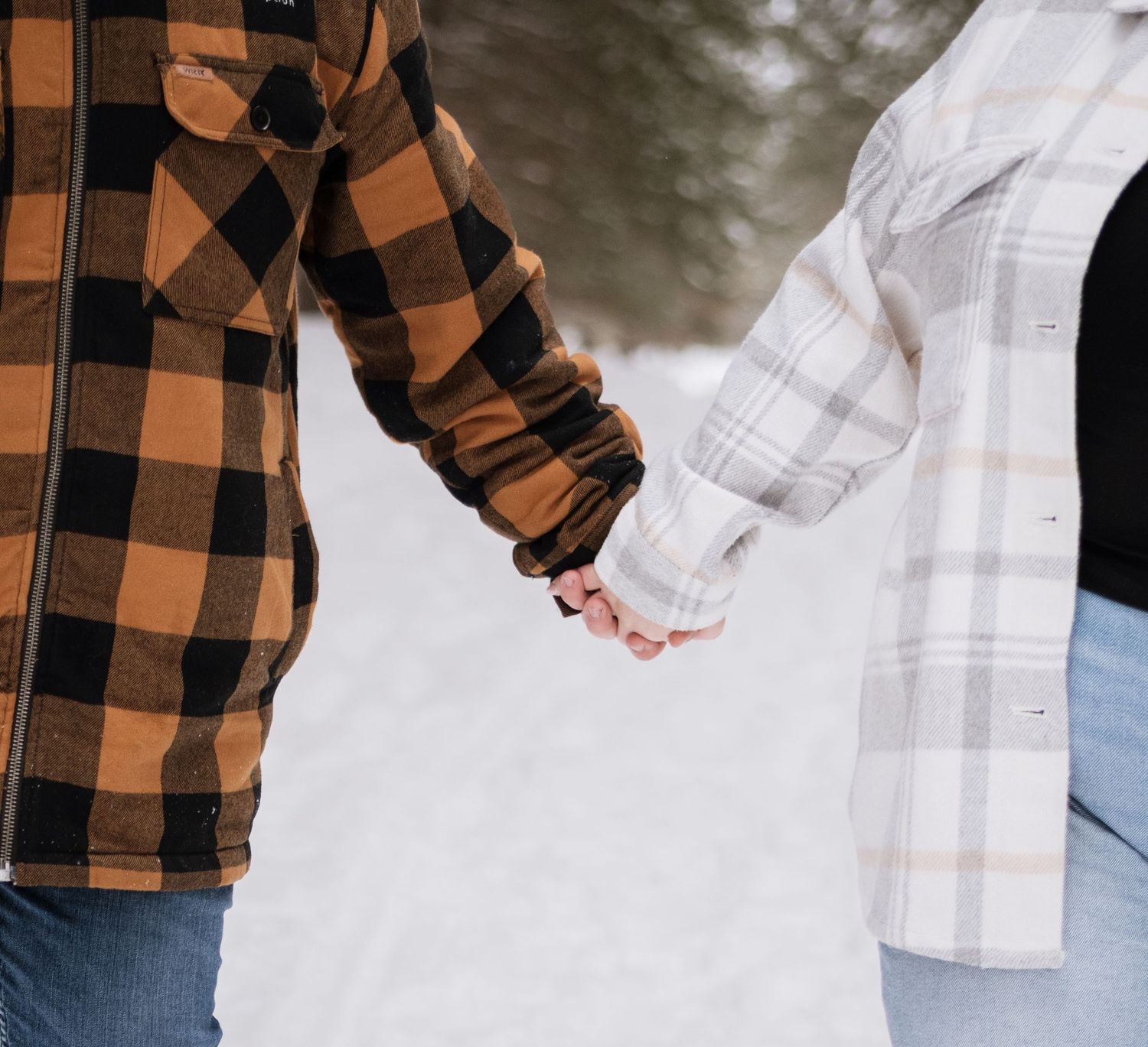 Close-up of couple holding hands in snow, man in orange buffalo plaid and woman in white plaid flannel shirt walking together.