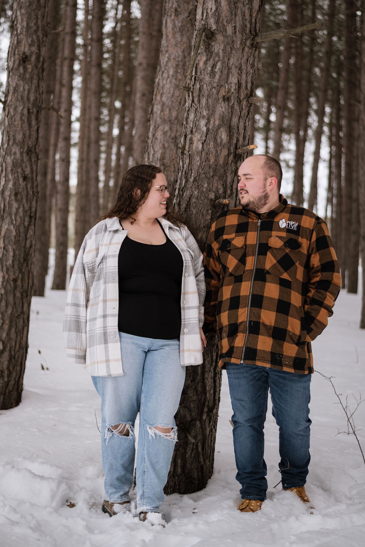 Couple in flannel shirts sharing a loving gaze beside a pine tree in a snowy winter forest.