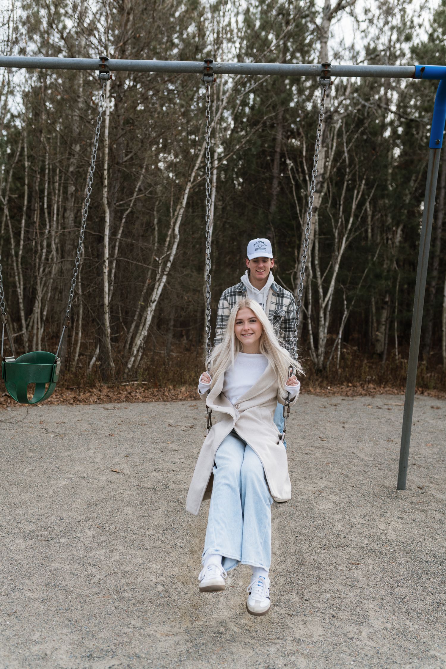 A couple at a playground, woman sitting on swing while man stands behind her pushing, surrounded by bare trees.