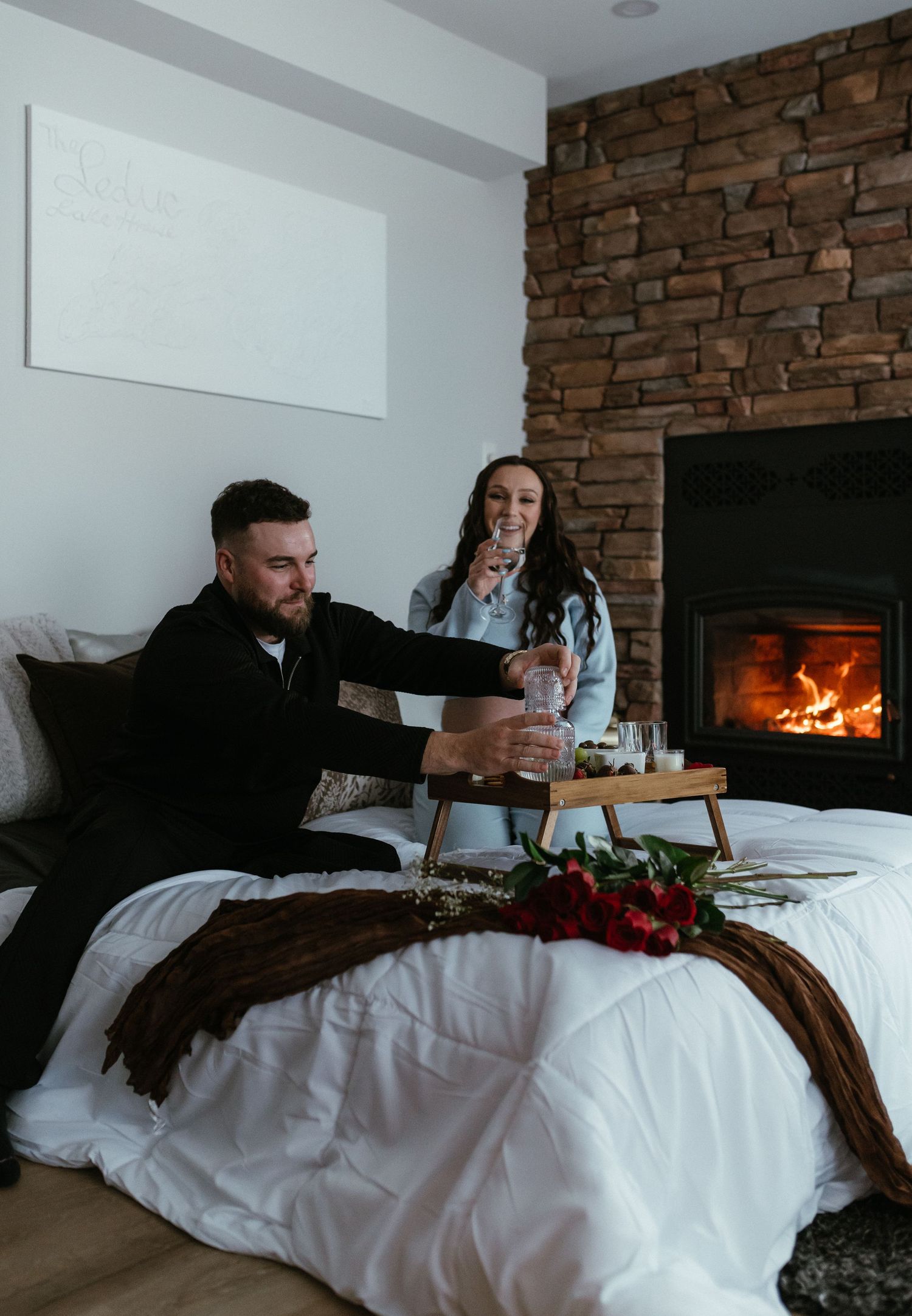 Couple toasting drinks on a white bed beside a stone fireplace with red roses.