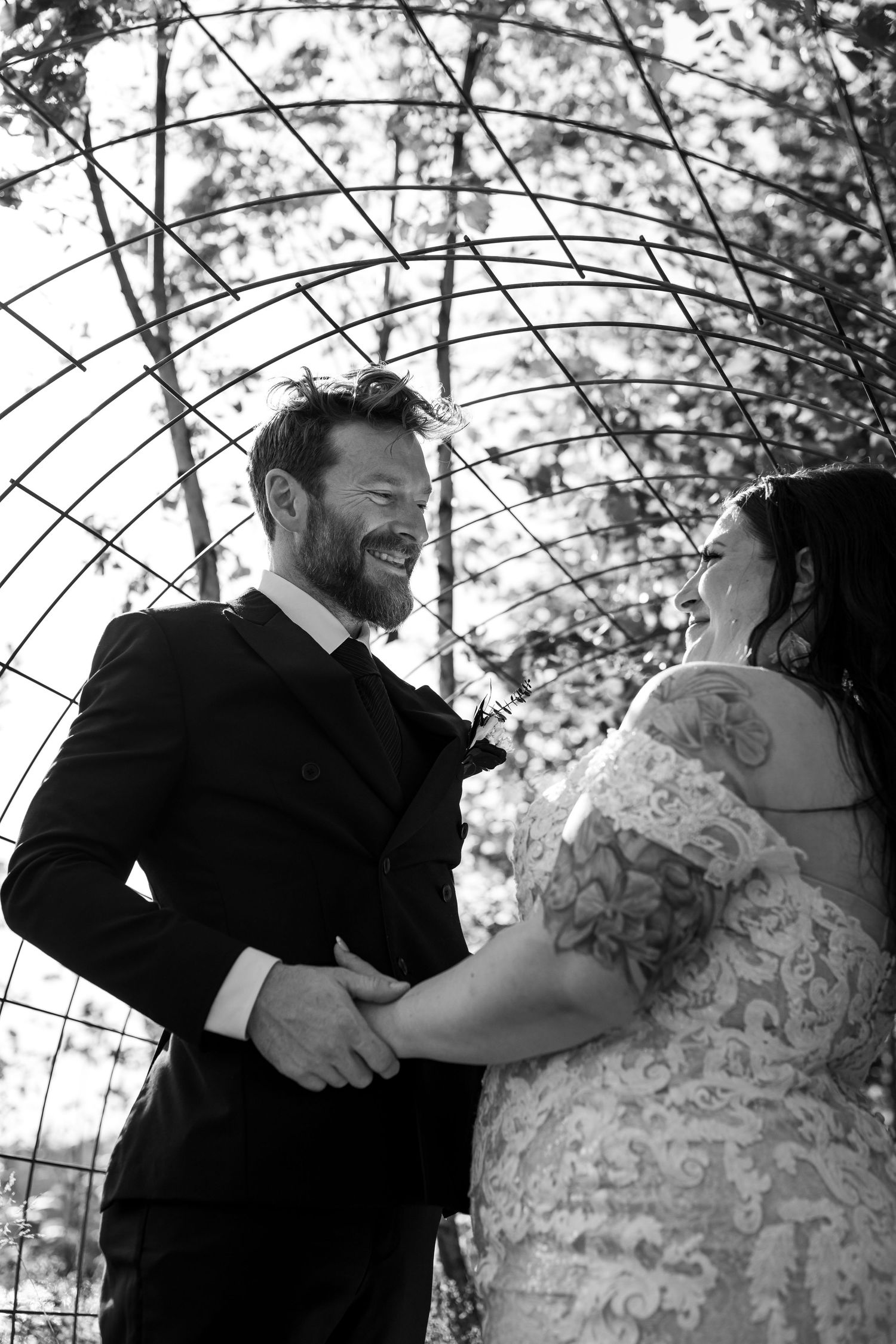 Black and white photo of a laughing groom in a suit holding hands with his bride in lace dress outdoors.