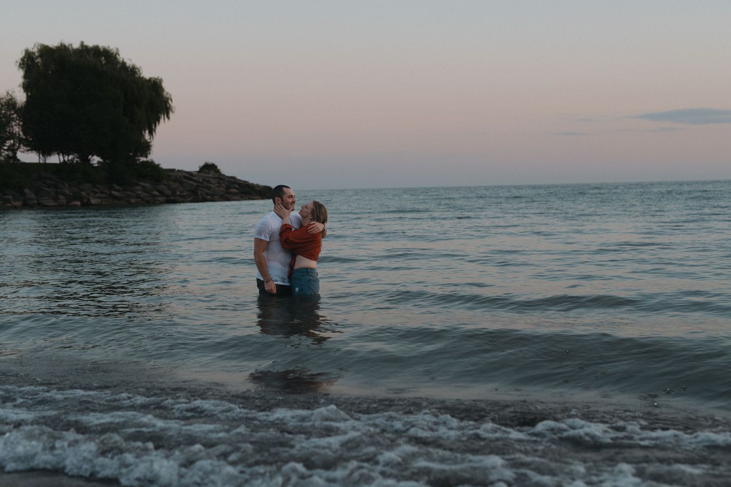 A couple shares an intimate moment while standing in ocean waves at dusk.