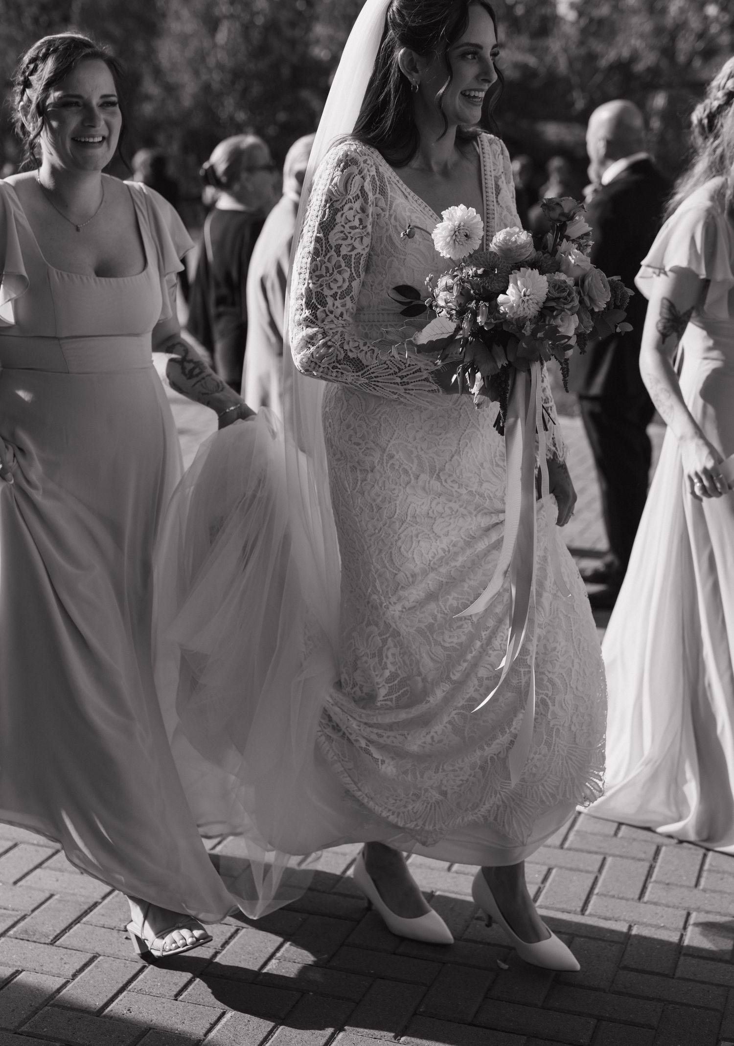 Bride in lace gown with bridesmaids walking on brick pavement in black and white.