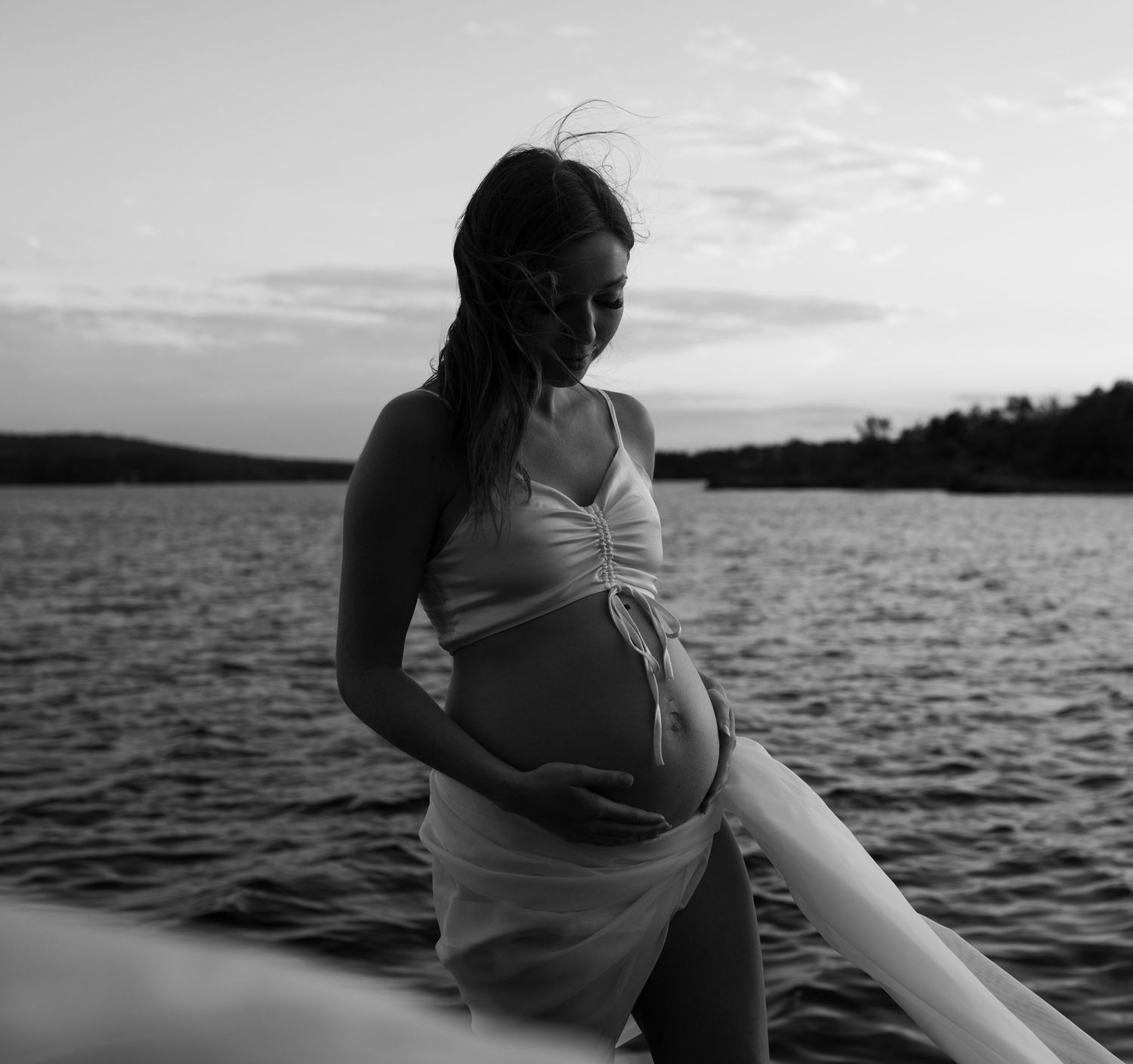 Silhouetted figure in white dress stands by calm lake at sunset creating a serene maternity photoshoot scene.