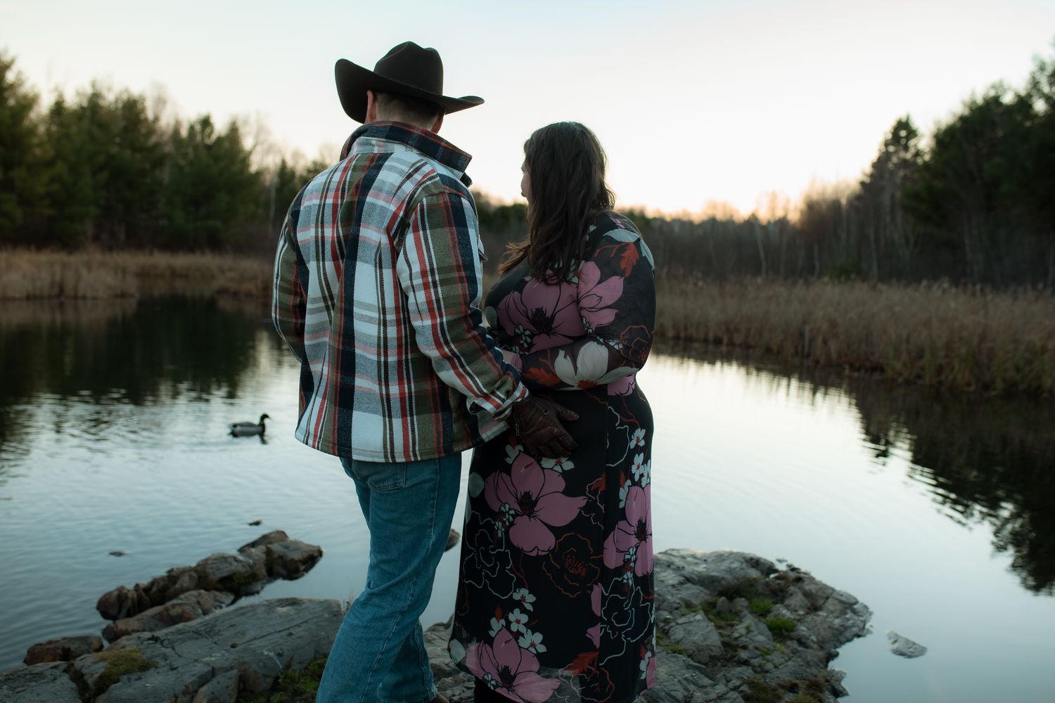 A couple stands by a serene lake at sunset, silhouetted against the tranquil water and trees.