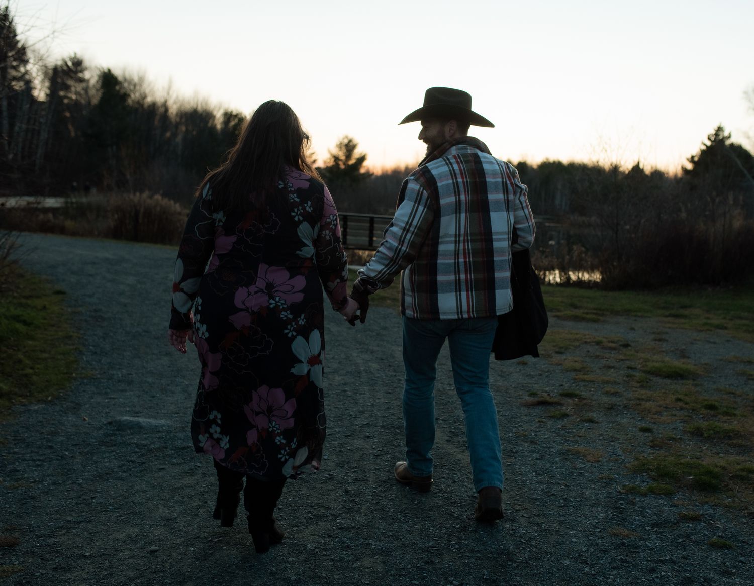 People walking along a dirt path at sunset with trees and fields in the background.