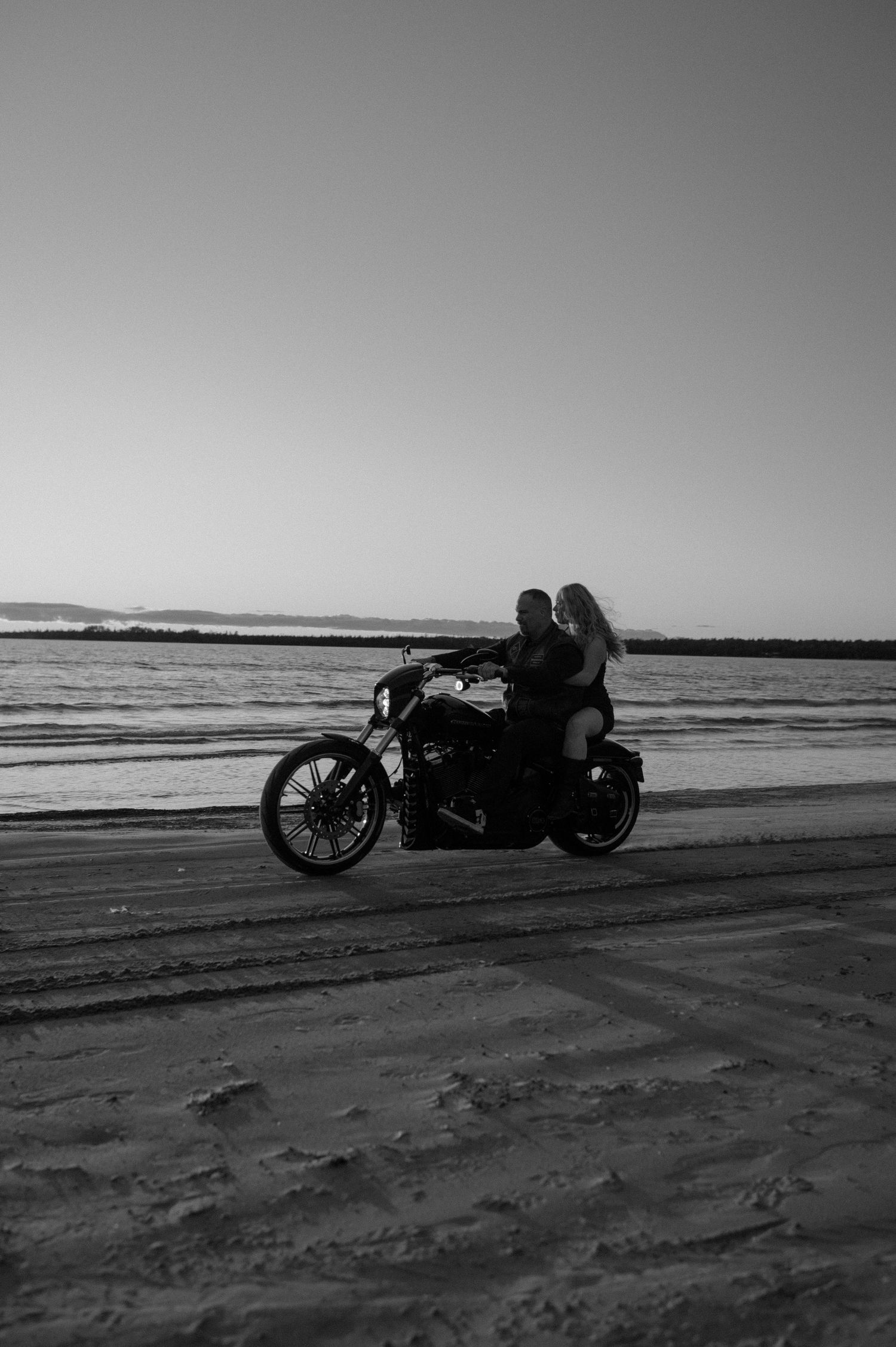 Black and white photo of a couple sitting on a motorcycle on a sandy beach at sunset near the ocean.