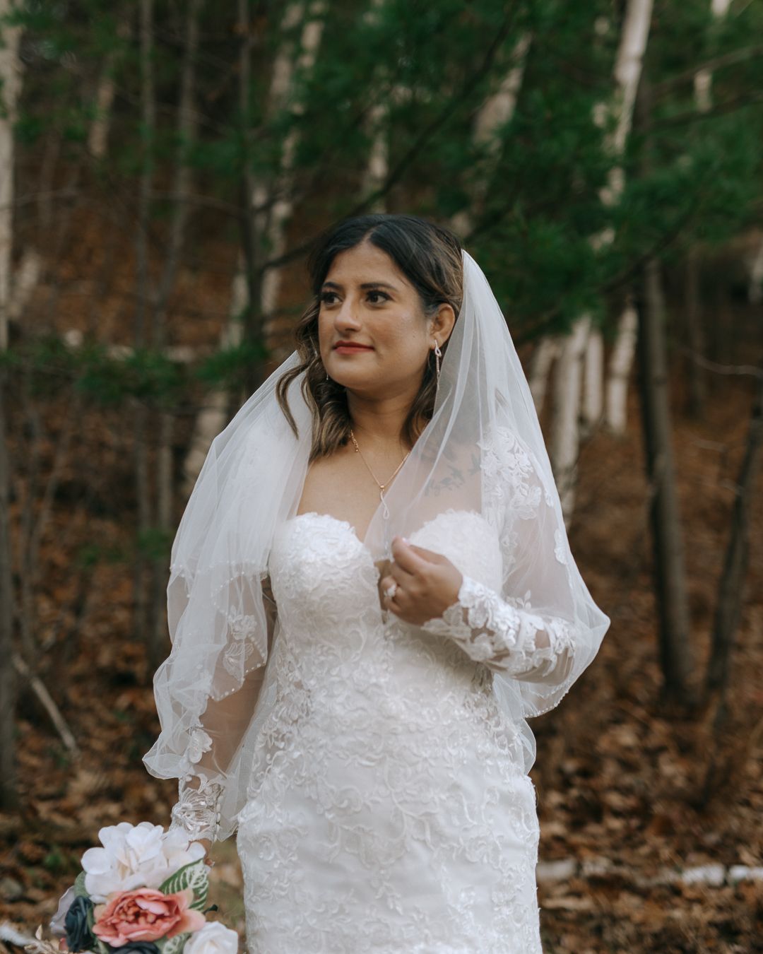 Elegant bride in white lace dress poses in autumn forest setting.