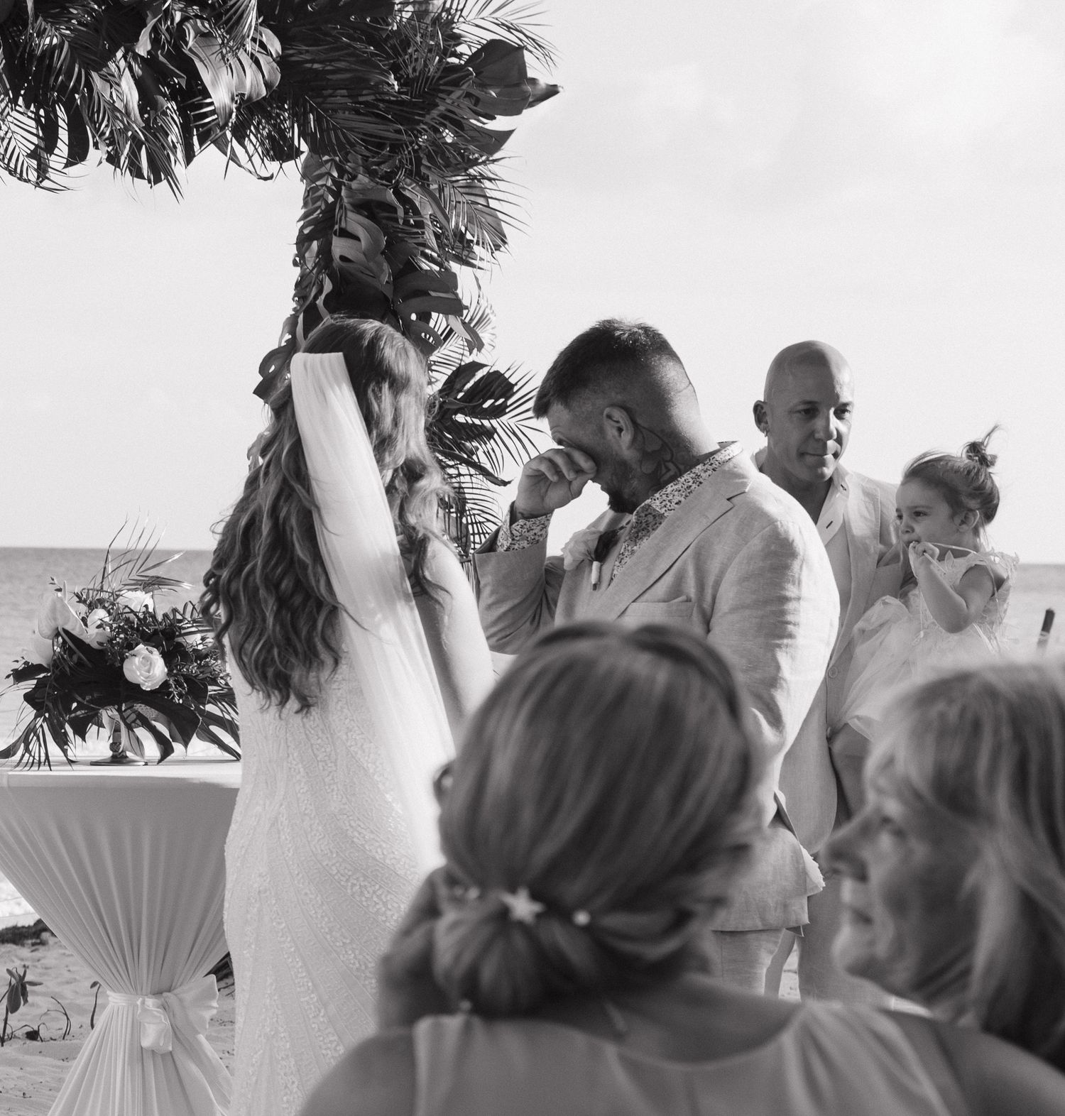 Black and white photo series of a beach wedding ceremony with palm trees and guests gathered on a sunny day.