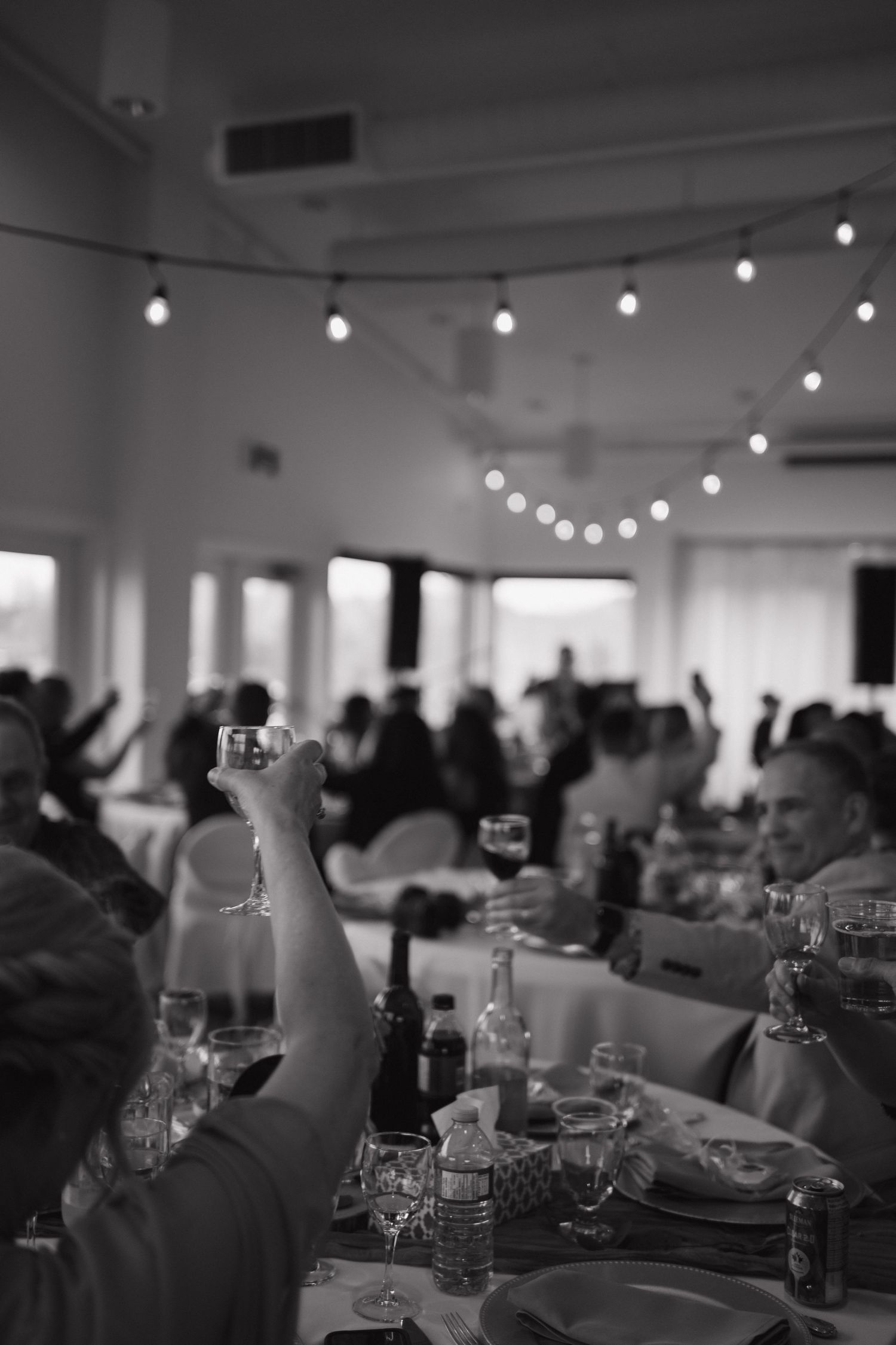 Black and white photo of wedding guests raising glasses in toast under string lights at reception.