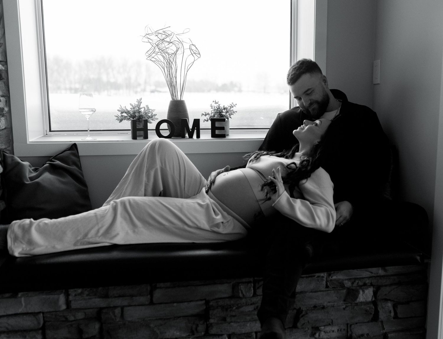 Black and white photo of a pregnant woman reclining on a window seat with her partner by a HOME sign.