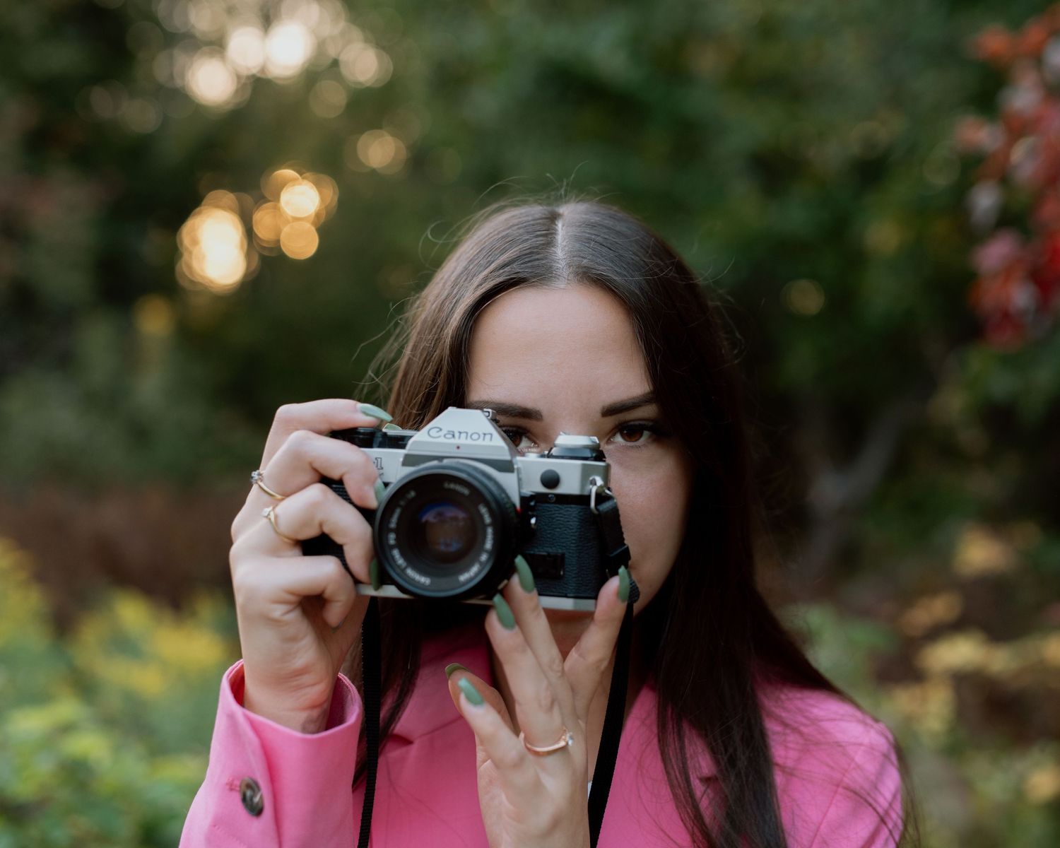 A young woman in a pink jacket holds a vintage film camera up to her eye outdoors in a green bokeh background.