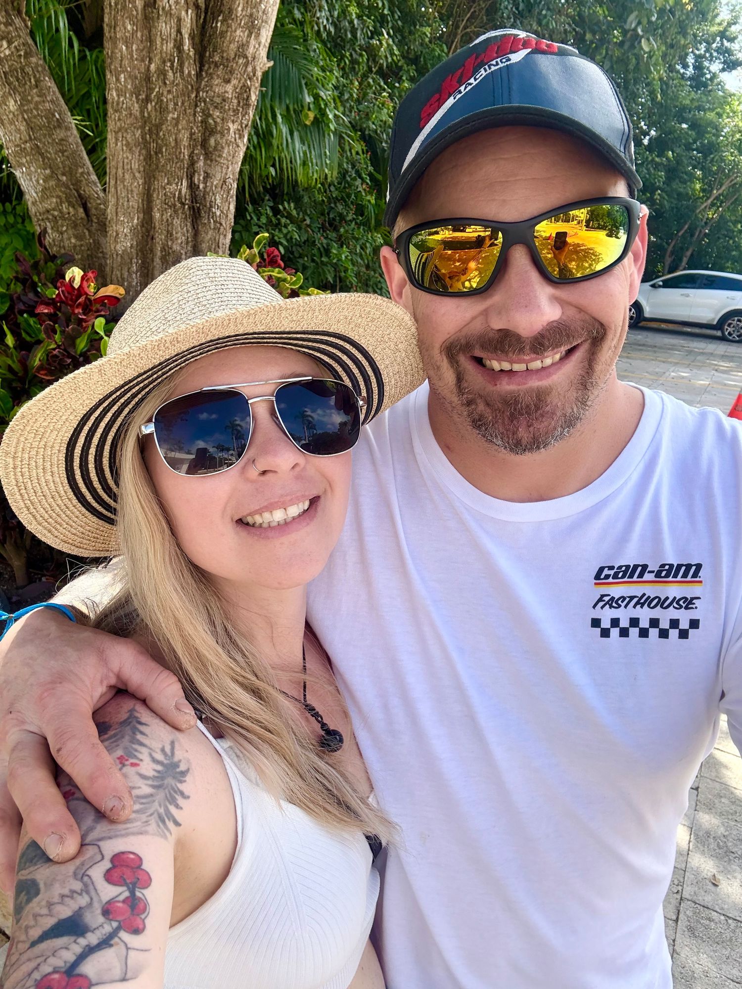 Smiling couple taking a selfie outdoors, woman in straw hat and sunglasses, man in Can-Am cap and white shirt.