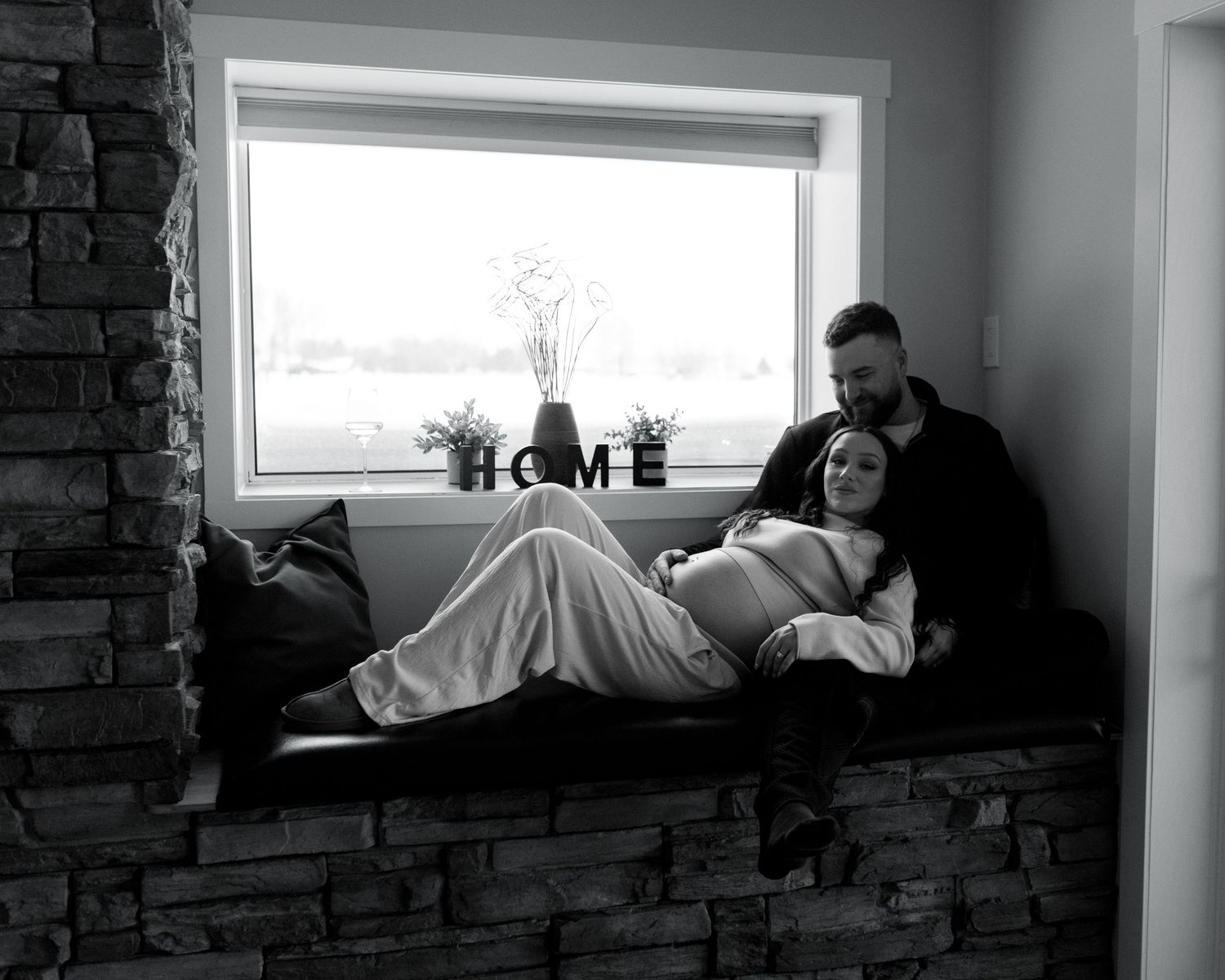 Black and white maternity photo of a couple cuddling on a window bench with a HOME sign nearby.