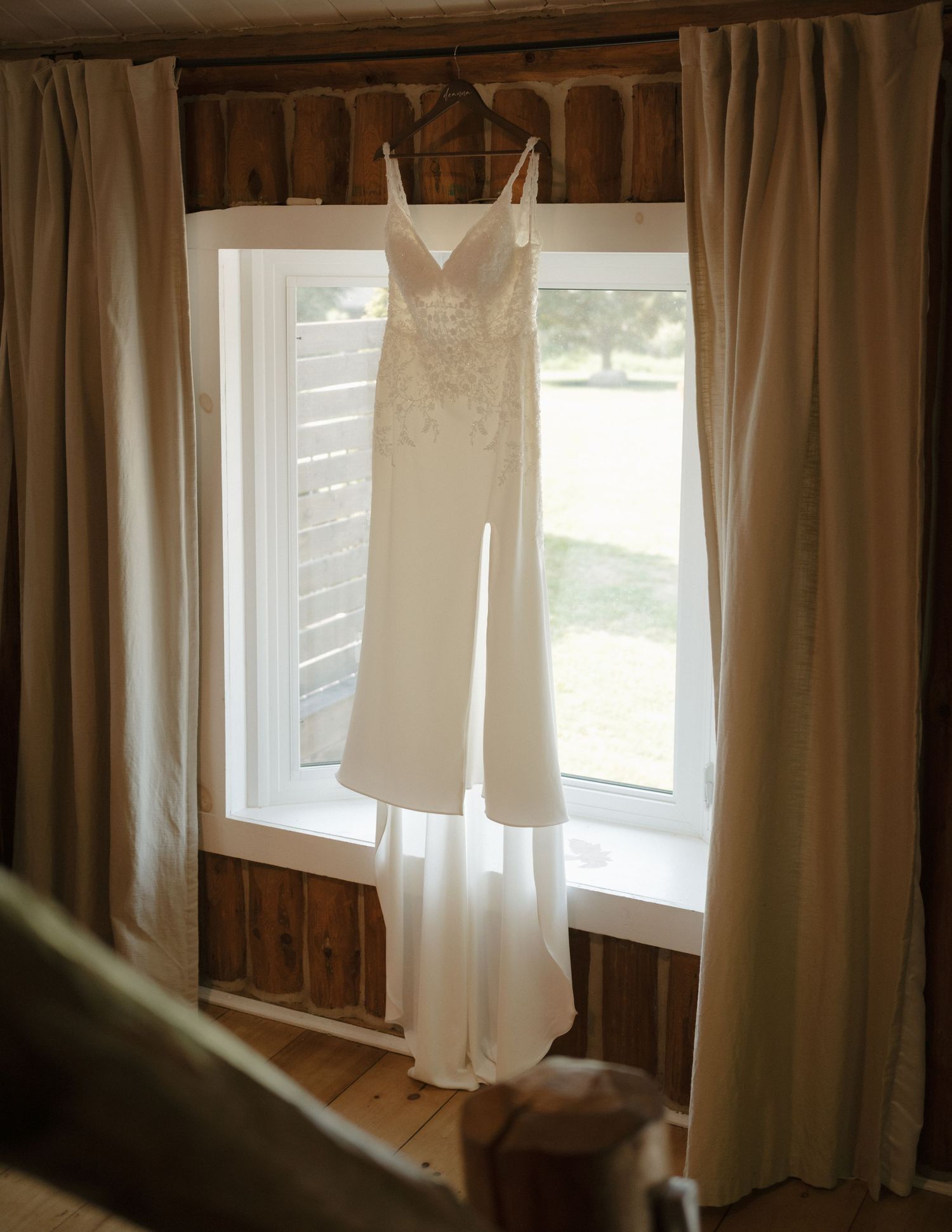 Elegant lace wedding dress hanging in a rustic barn window with cream curtains and natural light.