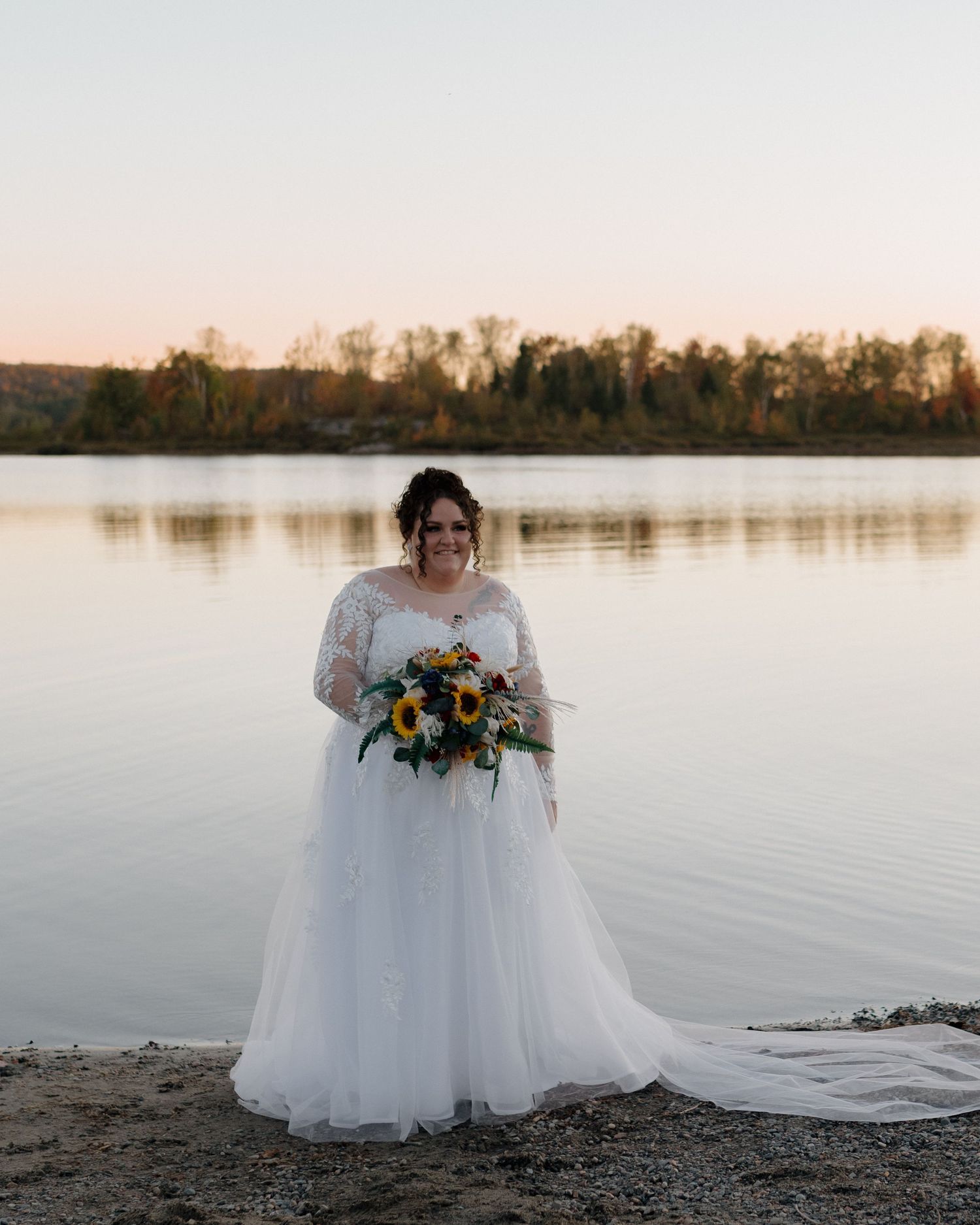 Bride in white lace long-sleeve gown holding sunflower bouquet standing by a calm lake at sunset.