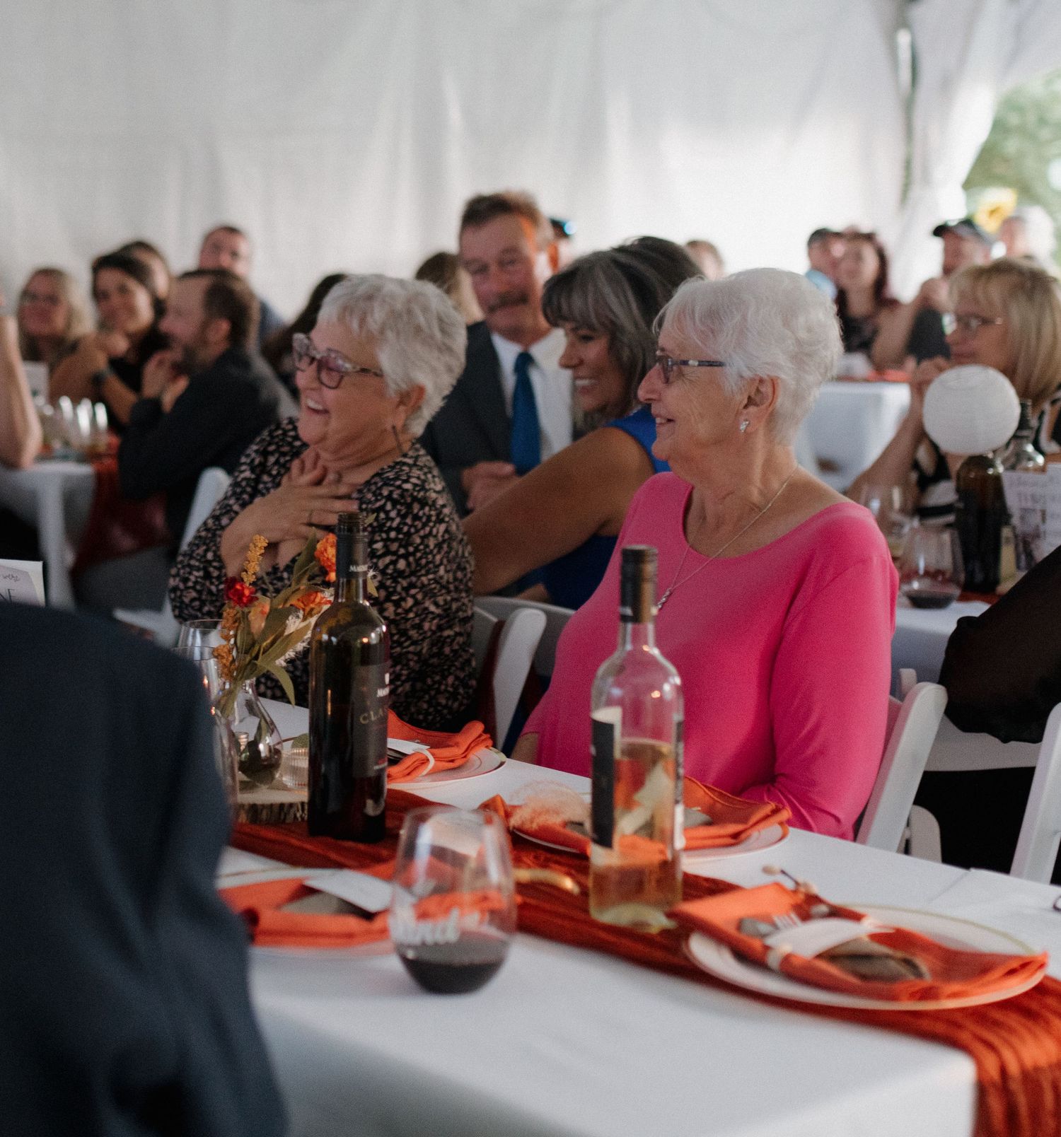 Wedding reception guests seated at tables with orange decor under white tent.