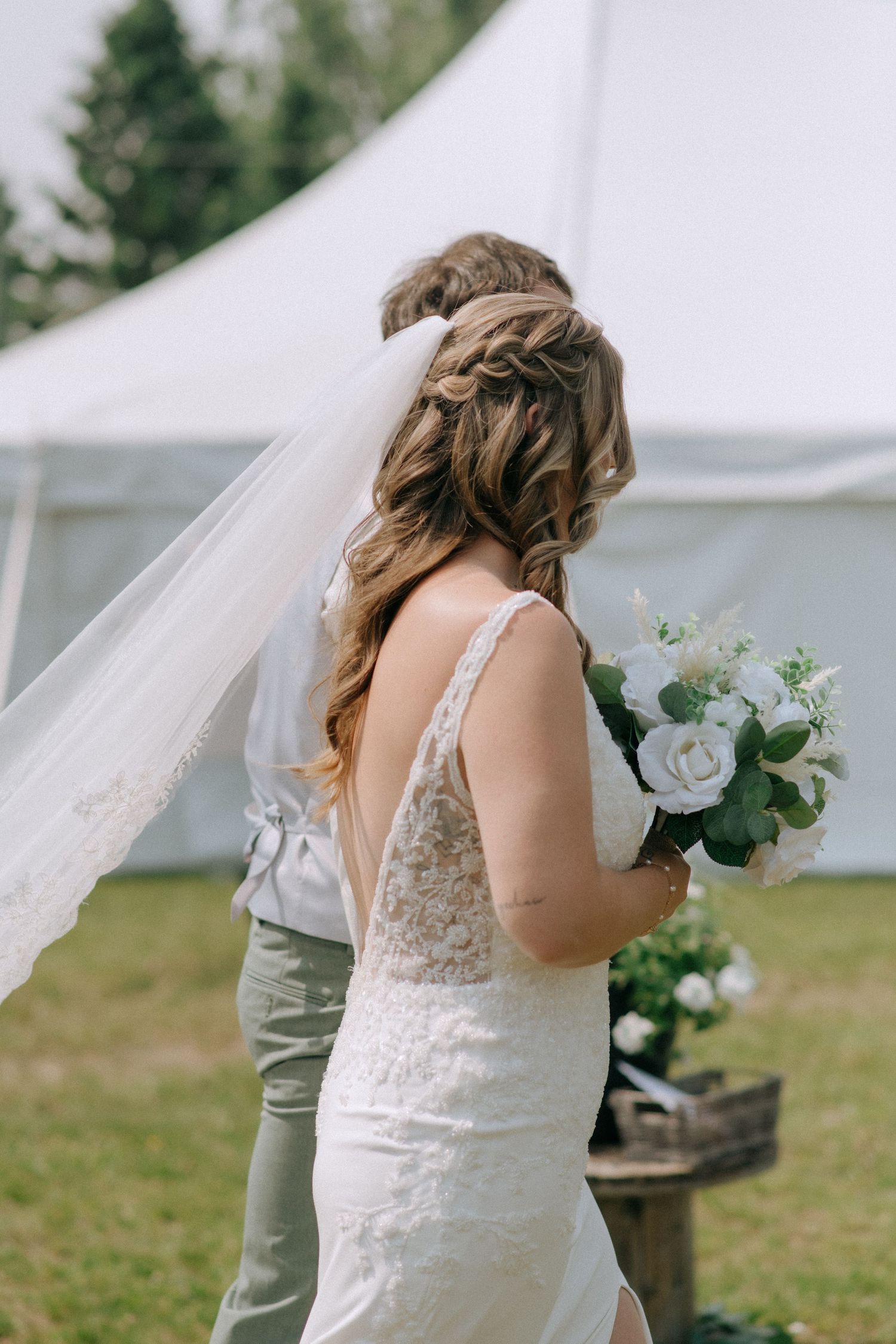 A bride in a detailed lace wedding dress holds a white floral bouquet next to a white tent outdoors.