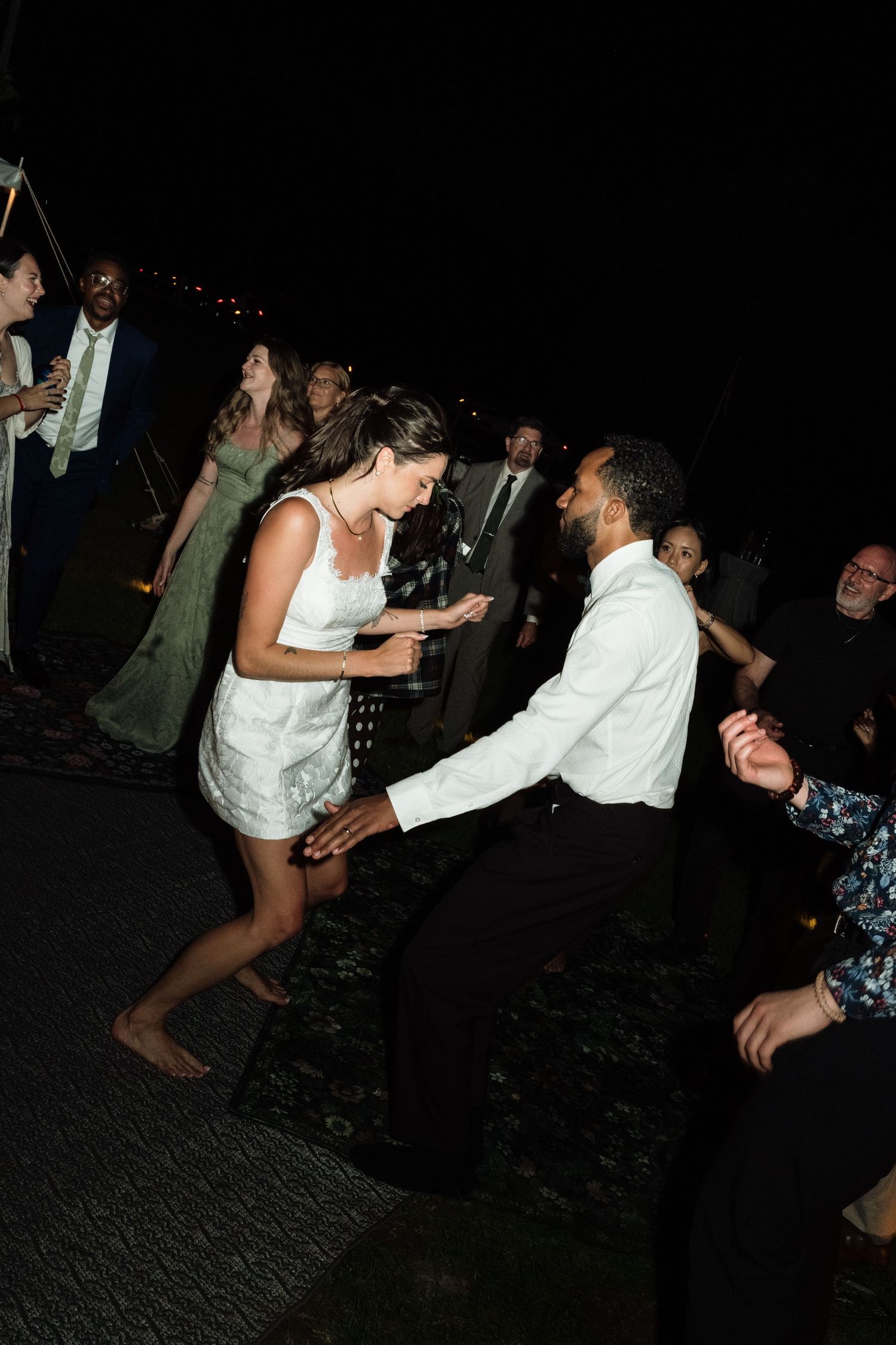 Bride and groom dancing joyfully at outdoor wedding reception at night with guests watching.
