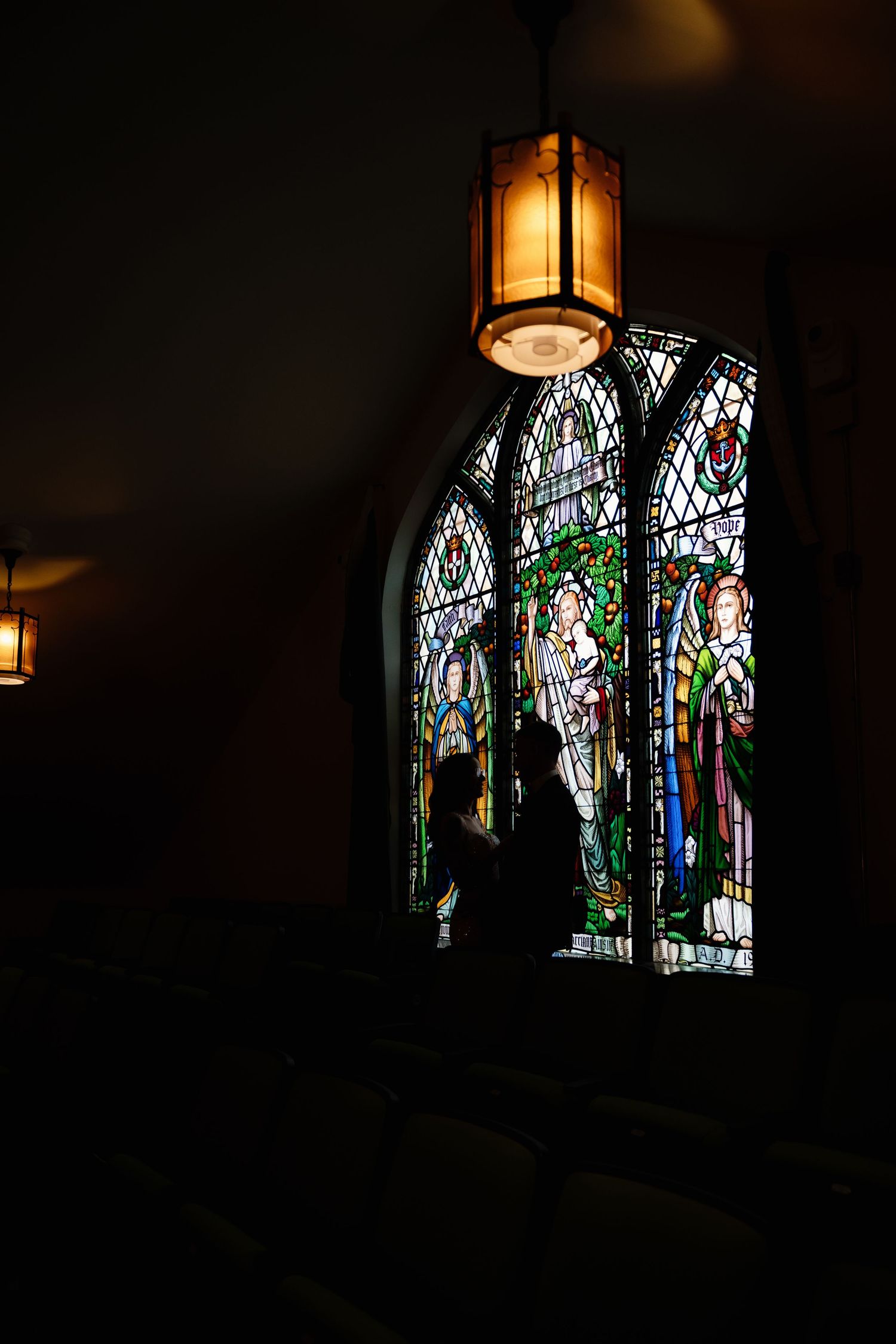 Illuminated stained glass window with religious figures in a dark church interior with hanging lantern.