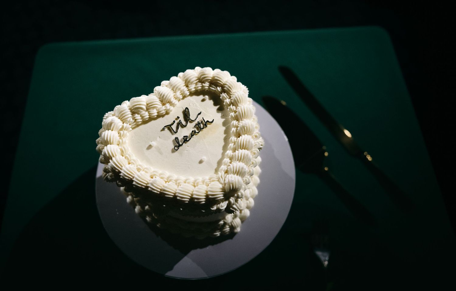 Heart-shaped white frosted Valentine's Day cake with decorative piping and inscription on a green surface.