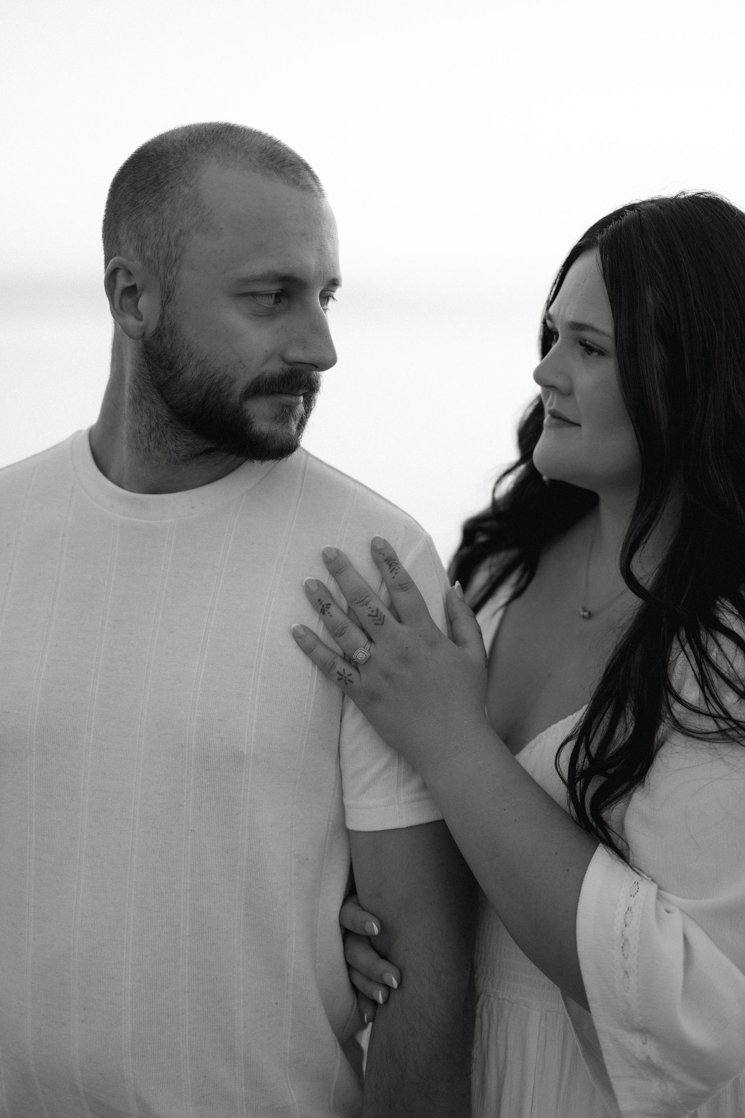Black and white photo of a couple, woman in white dress placing hand on mans chest, engagement ring visible.