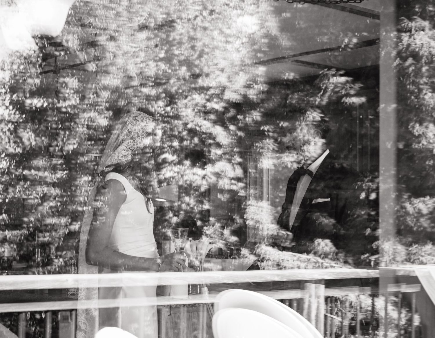 Black and white photo of a bride and groom on a reflective glass balcony surrounded by lush trees.