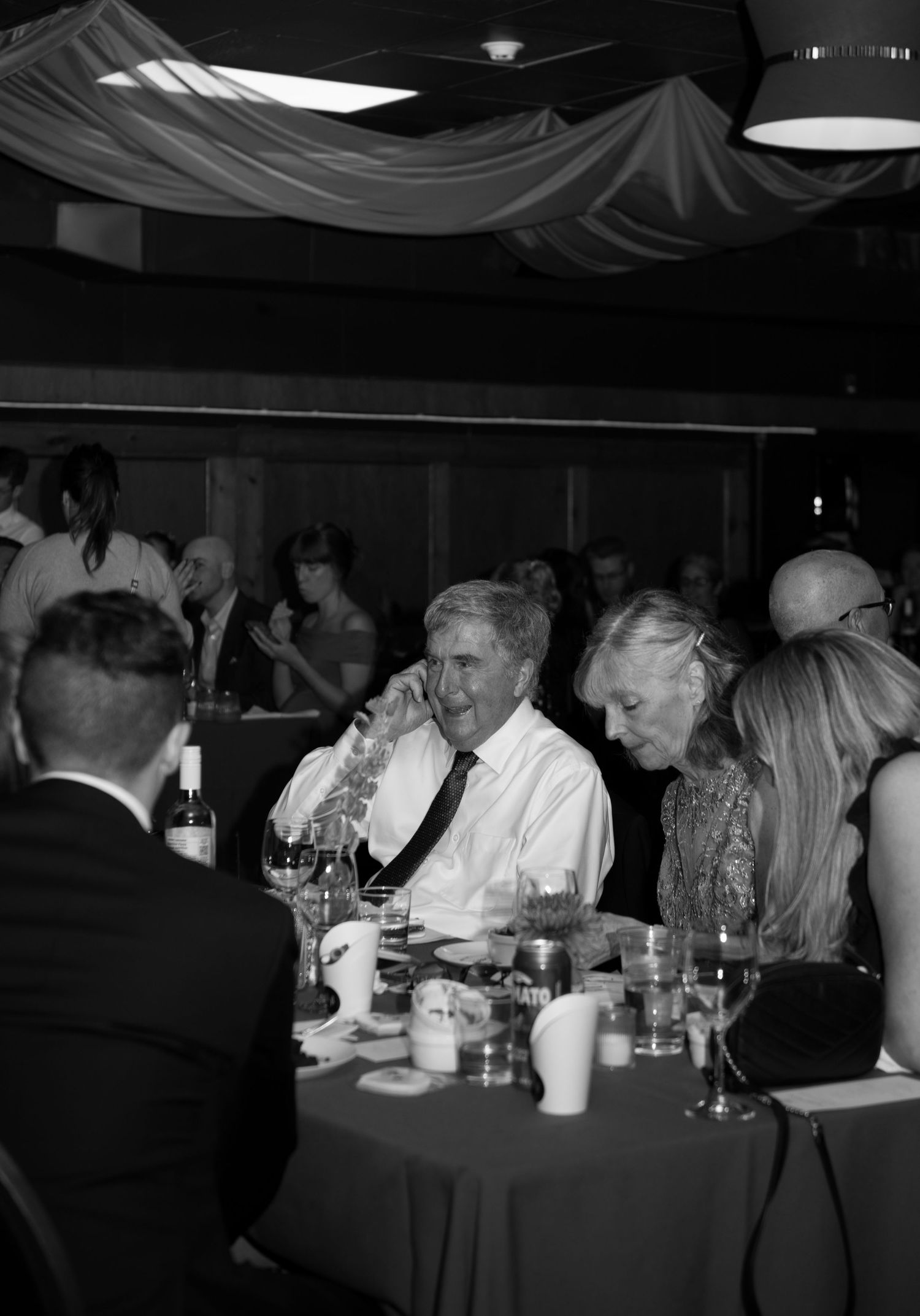Black and white photo of elegantly dressed guests dining at a formal event, with a man in a tie speaking at the table.