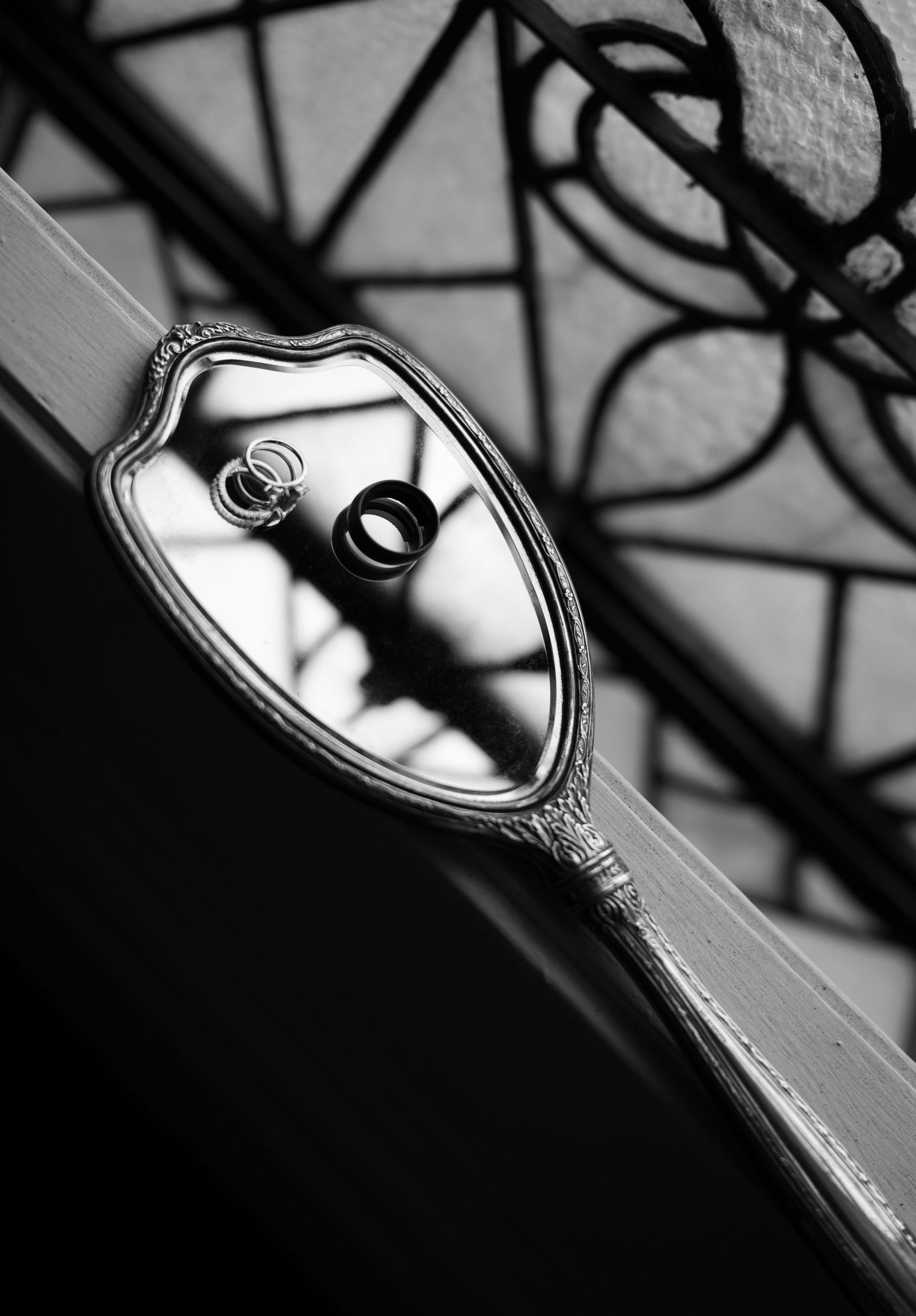 Black and white photo of wedding rings on a vintage hand mirror with ornate iron railing shadows.