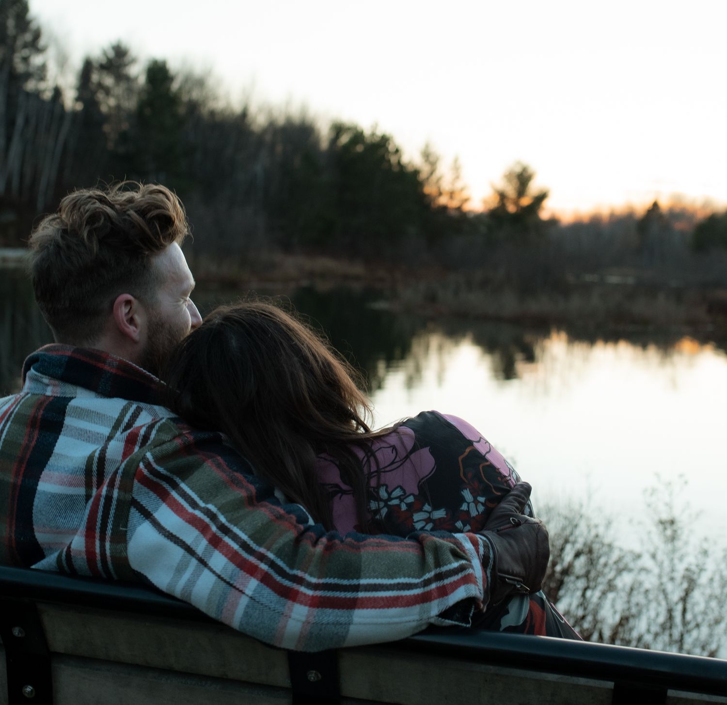 Silhouetted couple sits by tranquil lake at sunset, sharing an intimate moment against glowing sky and water.