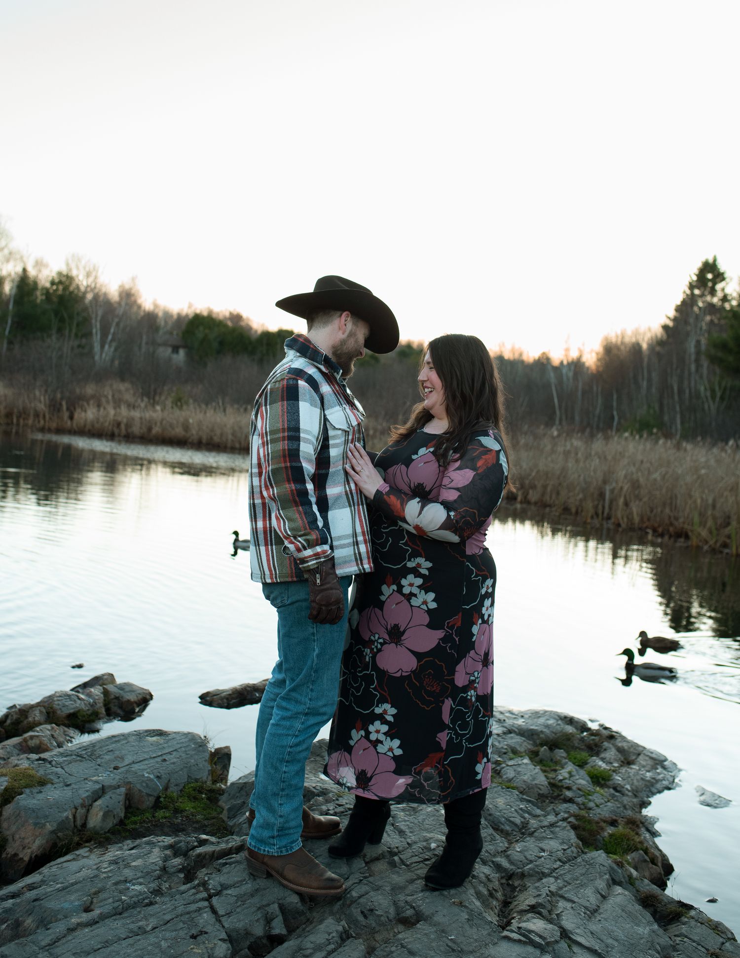 A couple stands together on rocks by a lake at sunset, with the person wearing a cowboy hat and plaid shirt.