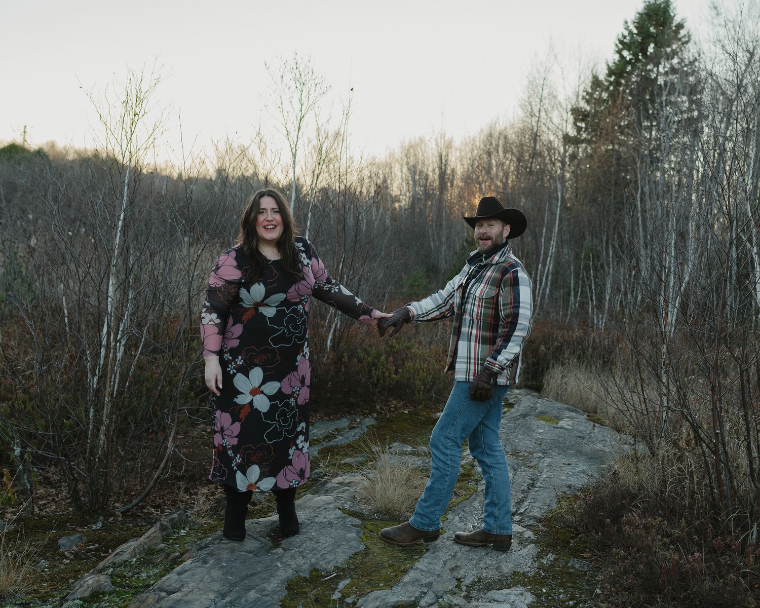 A couple strolls along a scenic forest trail during sunset, surrounded by bare winter trees and natural scenery.