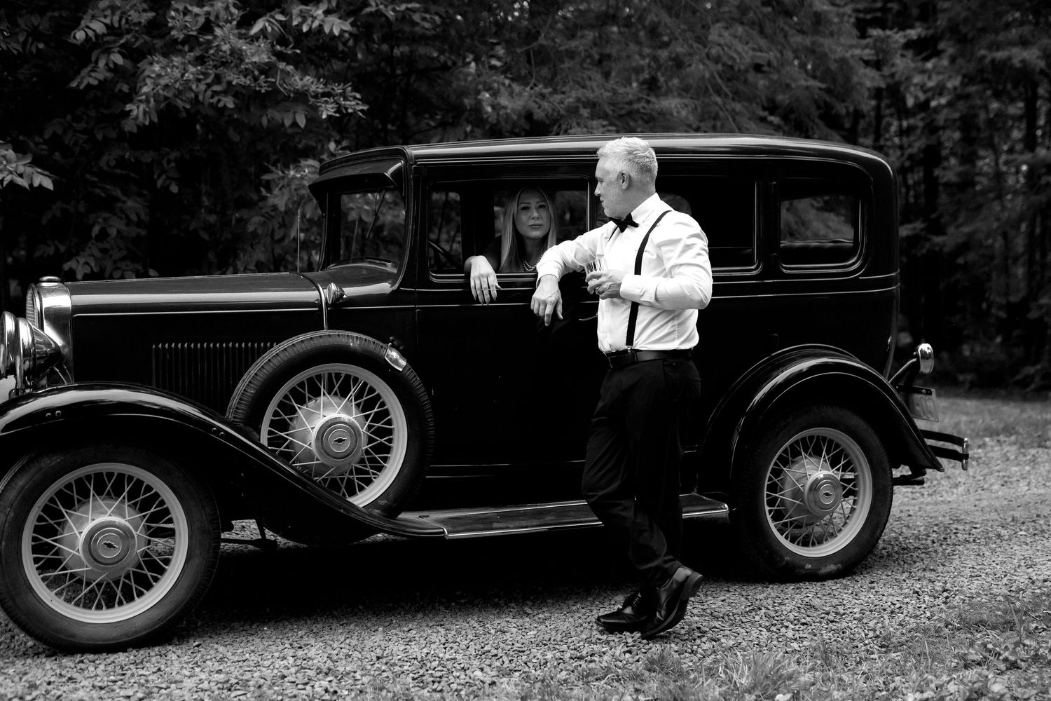Black and white photo of a couple posing beside a vintage classic car outdoors.