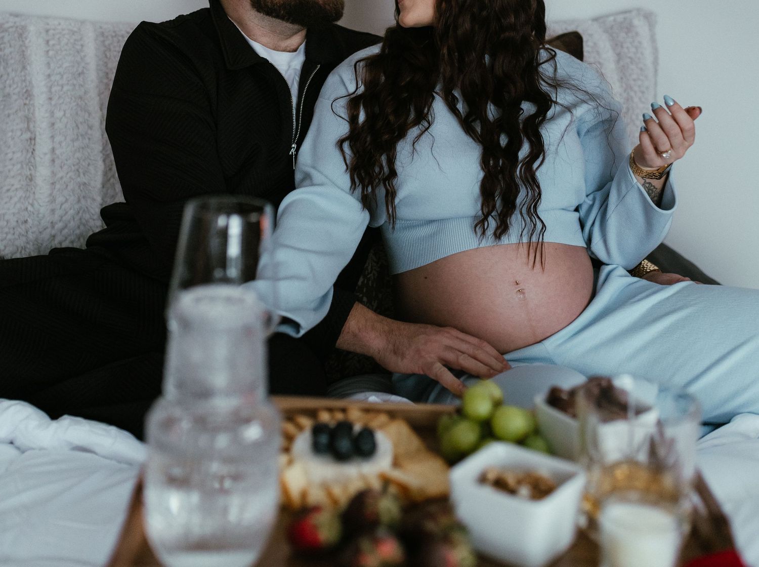 Pregnant couple sitting on bed with charcuterie board featuring fruits, nuts, and snacks in foreground.