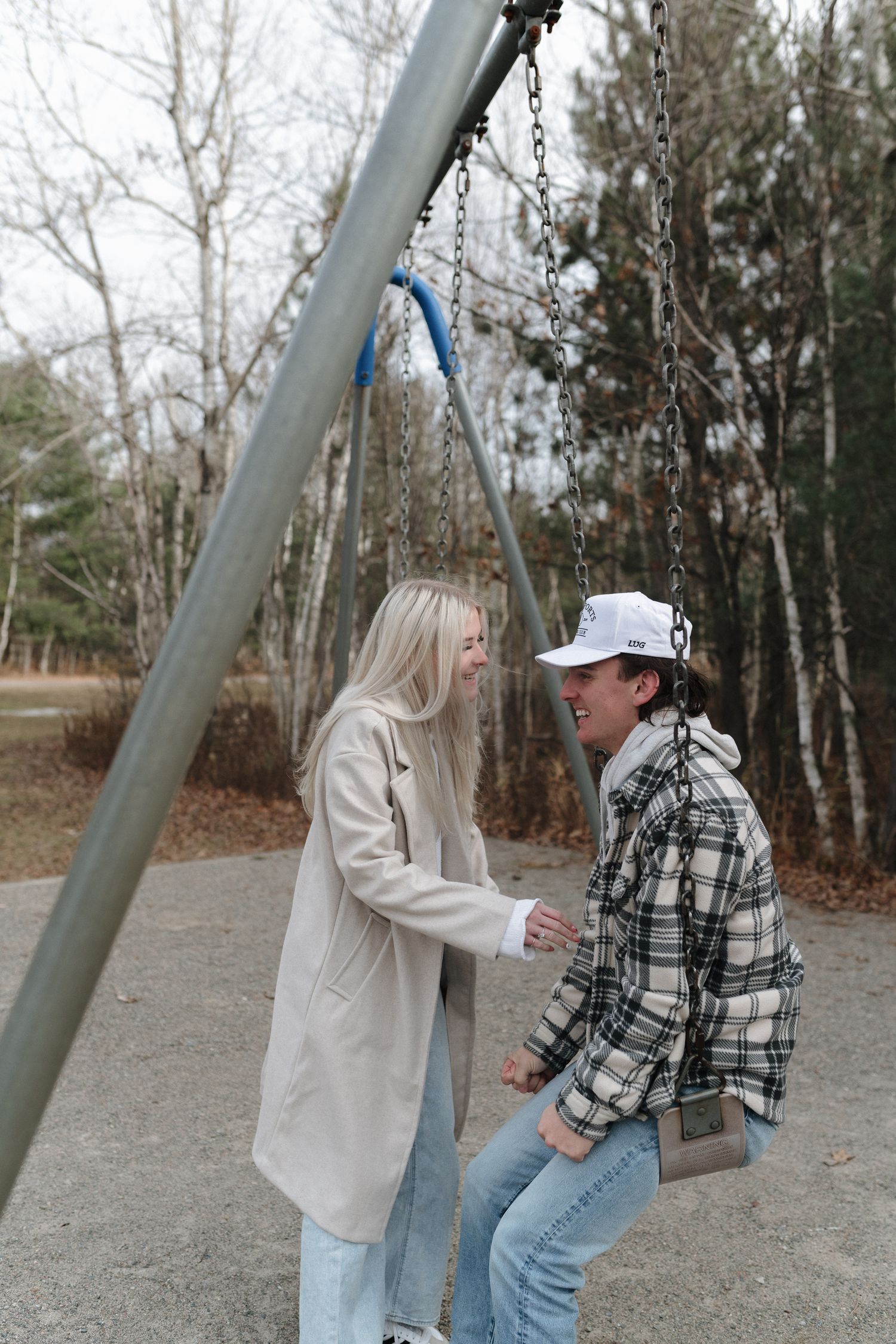 Couple laughing together at playground swing set surrounded by bare trees in fall.