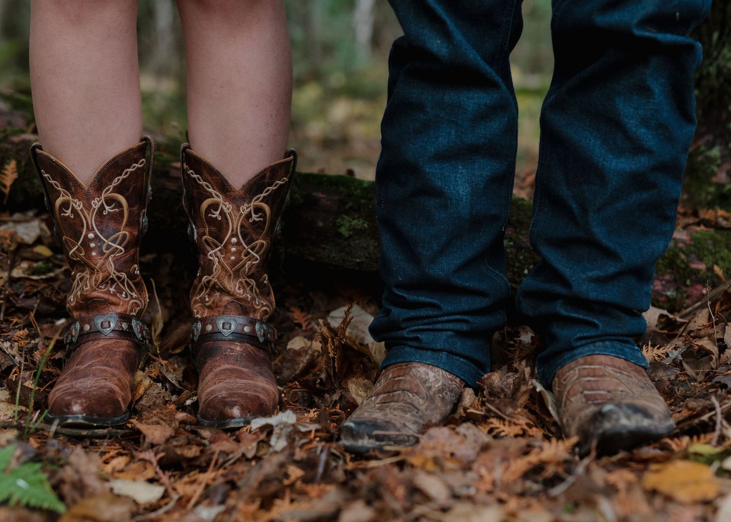 Close-up view of brown leather boots and blue jeans standing in autumn leaves on forest ground.
