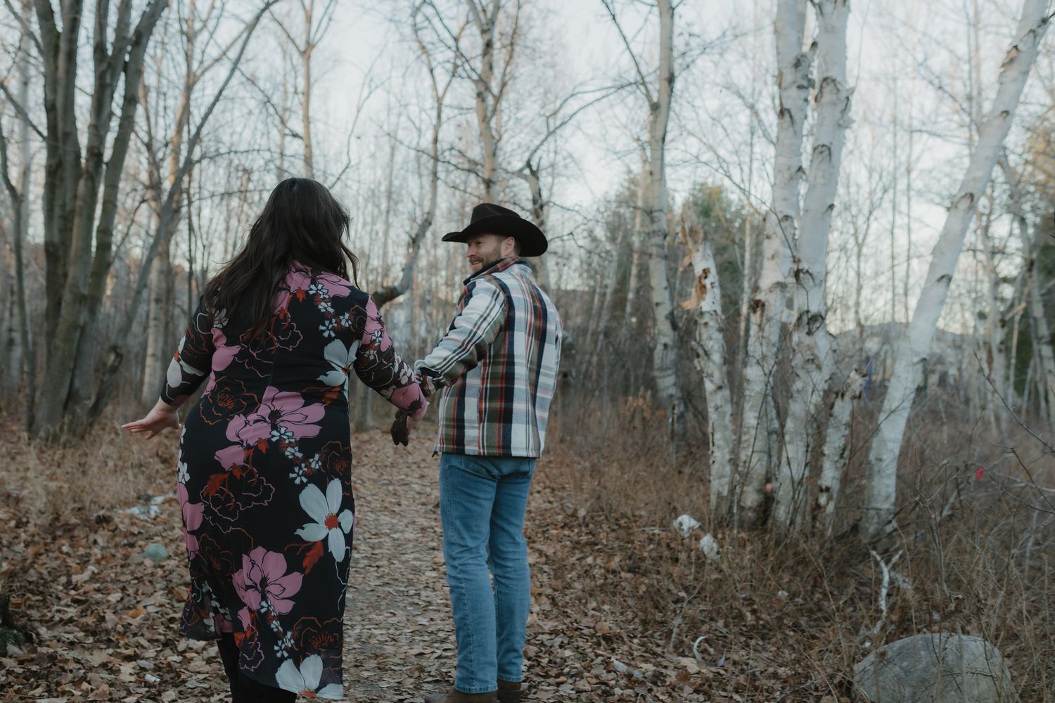 A couple walks hand in hand through a misty autumn forest with bare trees and fallen leaves on the ground.