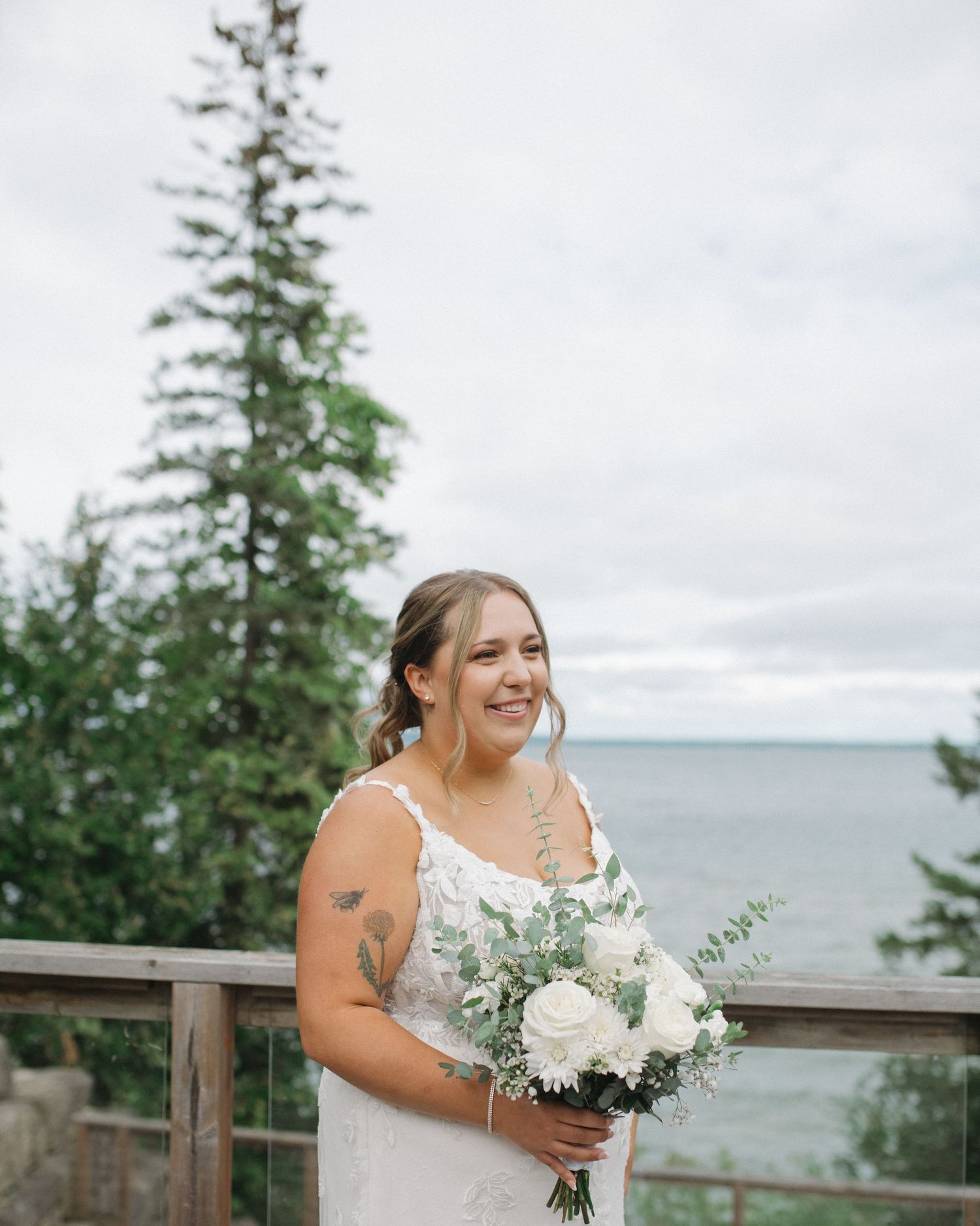 Bride holding white bouquet smiles joyfully on outdoor deck overlooking water.