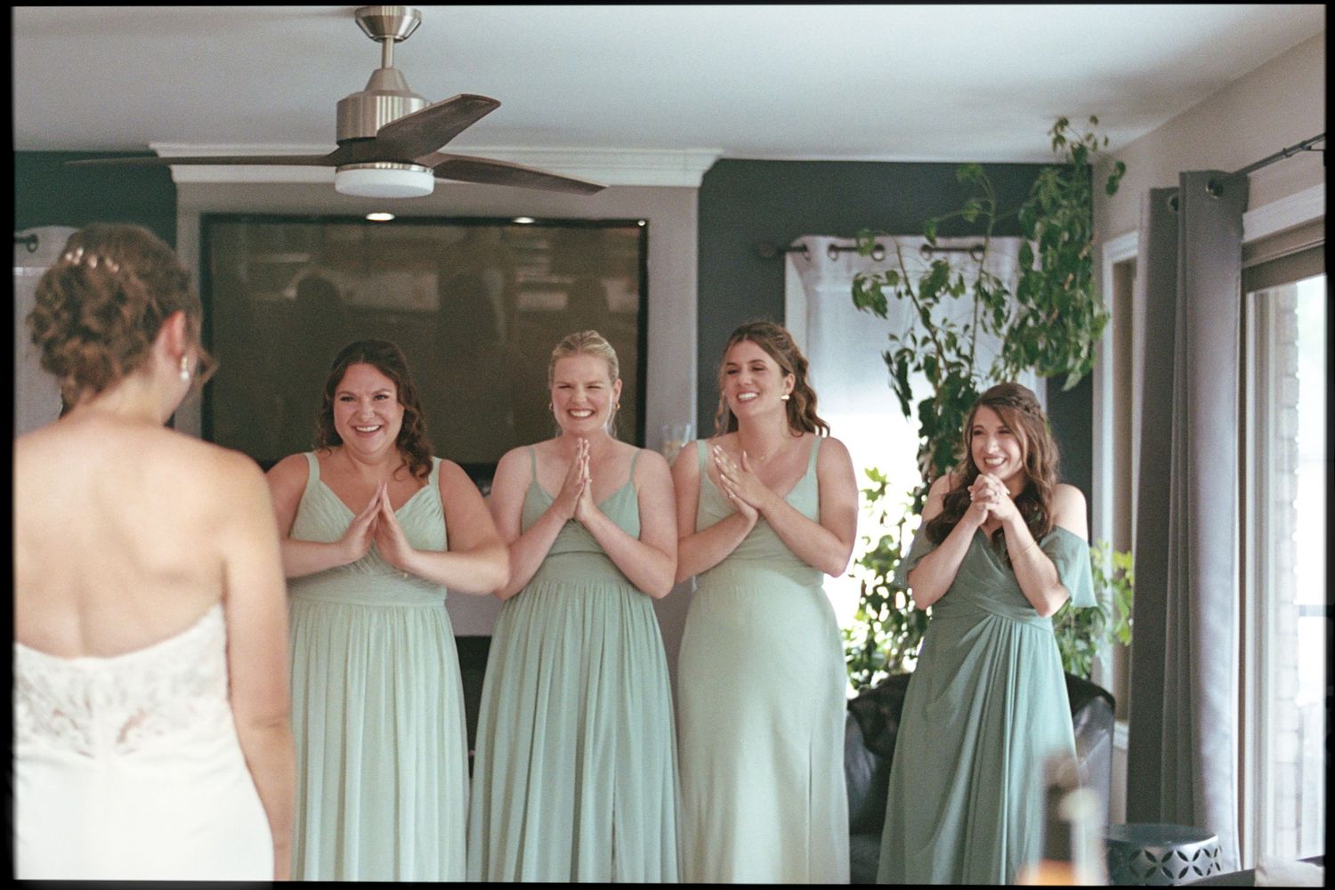 Bridesmaids in sage green dresses react with joy as bride reveals her wedding gown during bridal party preparations.