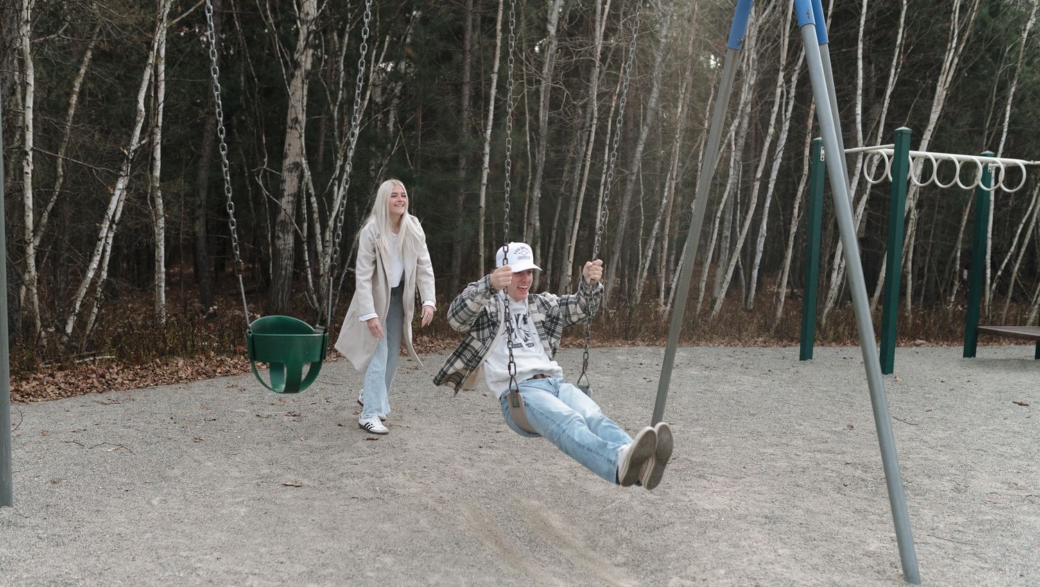Woman pushing a child on a swing at a playground surrounded by tall trees in autumn.