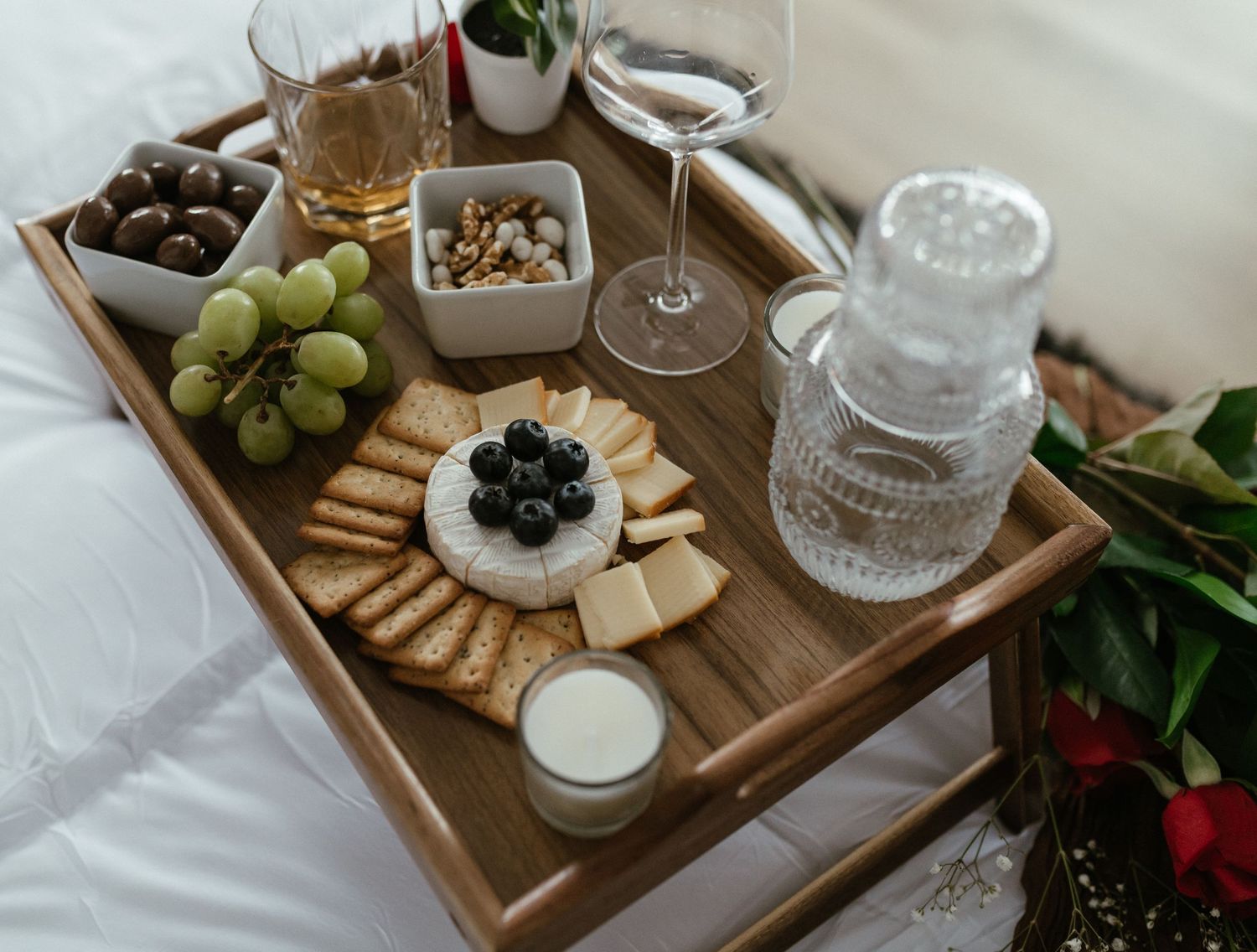 Romantic charcuterie board with cheese, crackers, grapes, and wine glasses on a wooden tray atop a white bed.