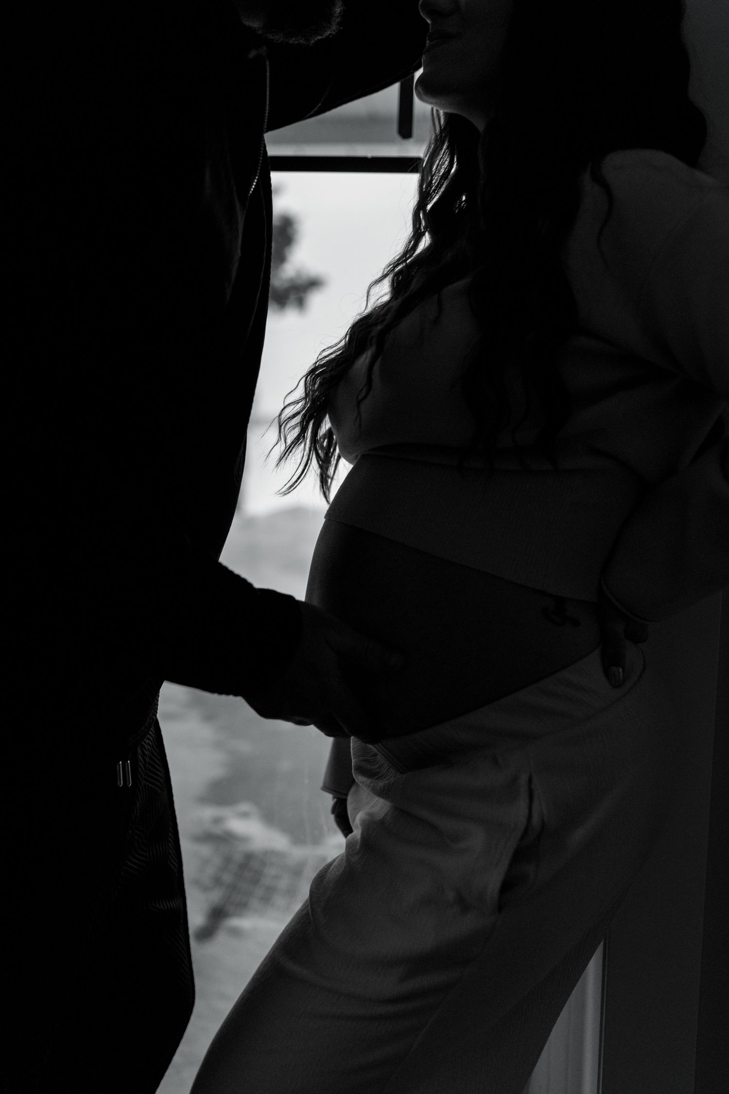 Silhouette of pregnant woman and partner standing by a bright window in black and white.