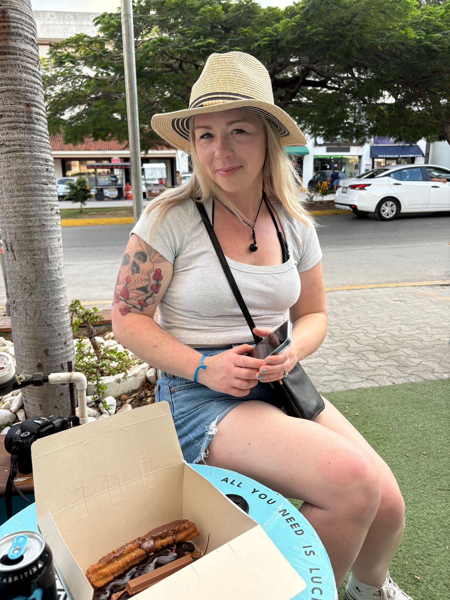 Blonde woman in straw hat and denim shorts sits outdoors with churros in a box, showing arm tattoo.