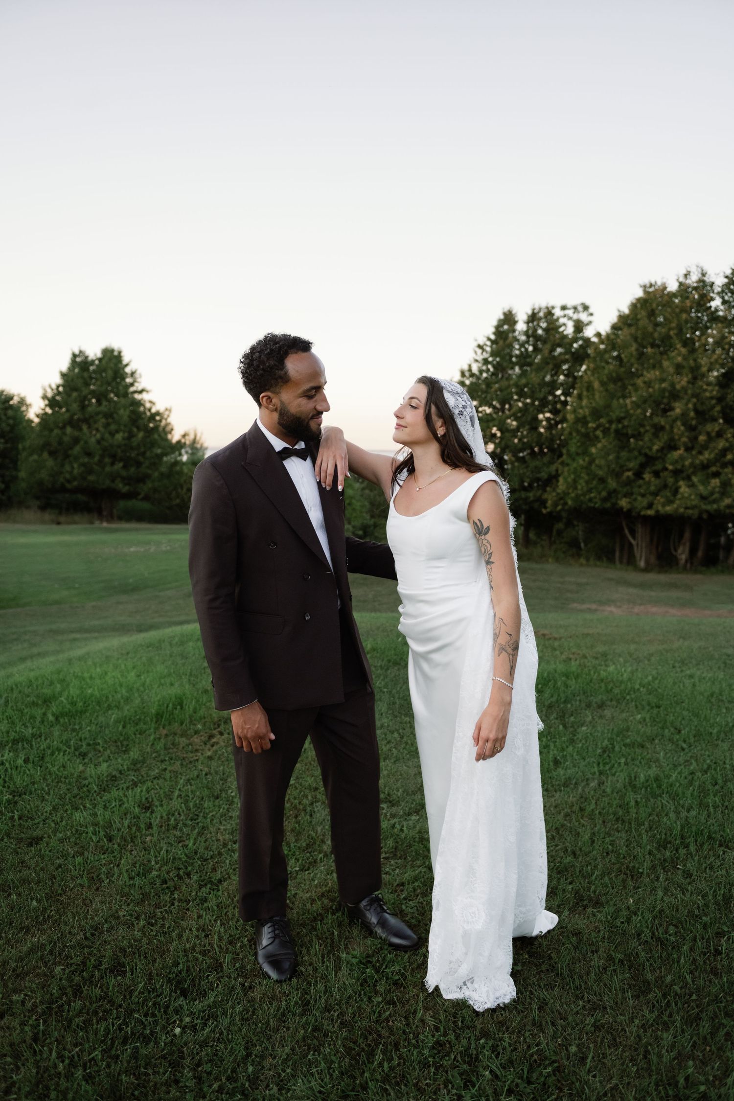 Interracial couple in wedding attire share a tender moment on a green lawn at dusk, bride in white gown with veil.