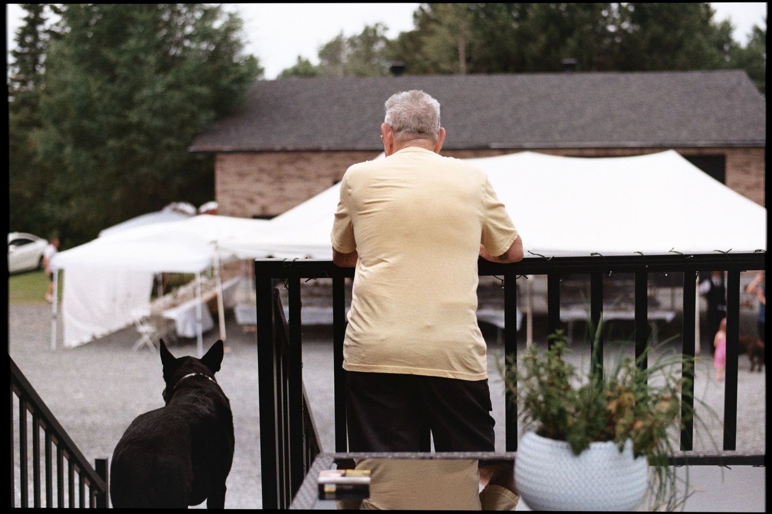 Elderly man in yellow shirt leaning on deck railing with black cat nearby, overlooking outdoor event tents.