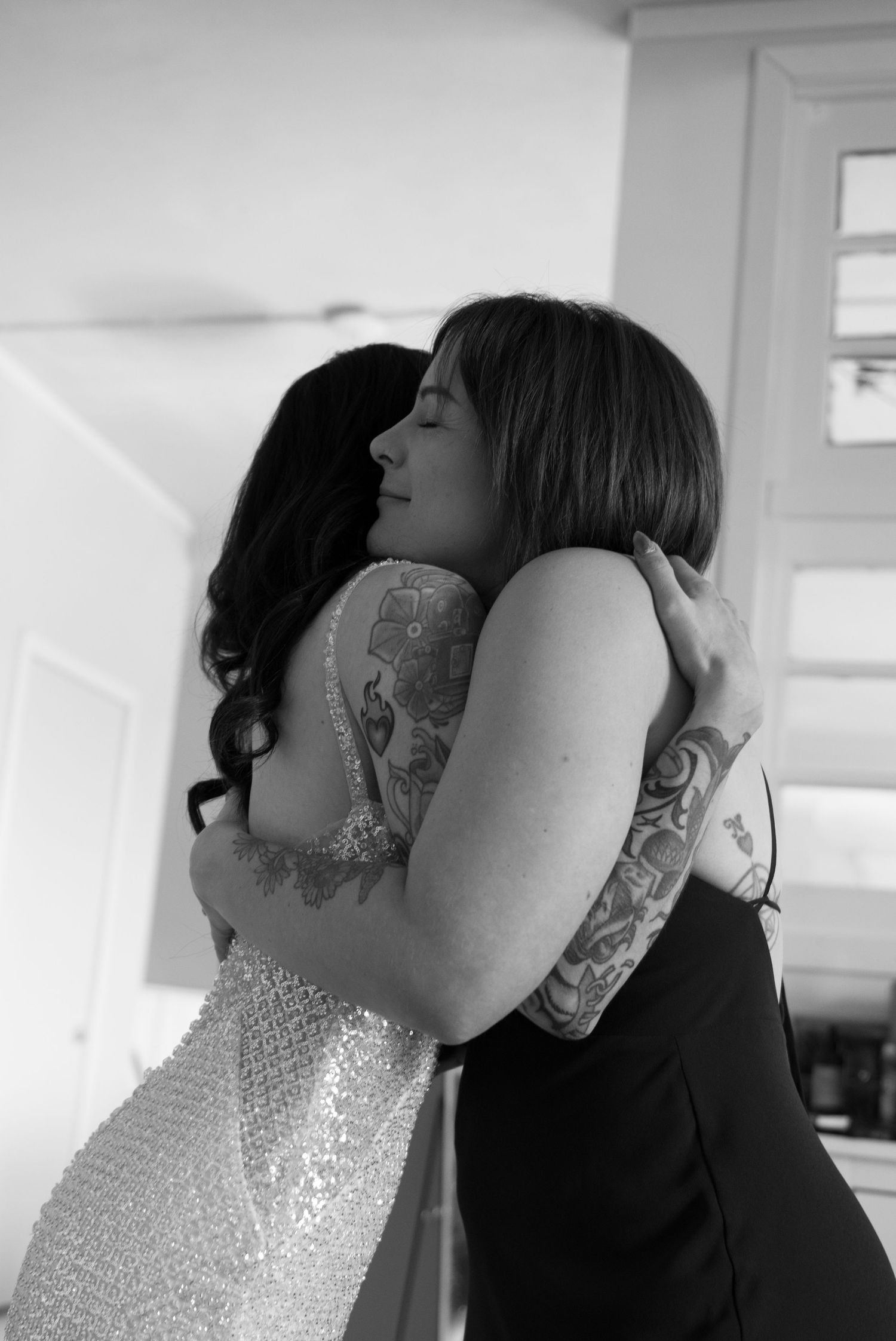 Two women embracing in a tender hug, one wearing a sequined dress, captured in black and white photography.